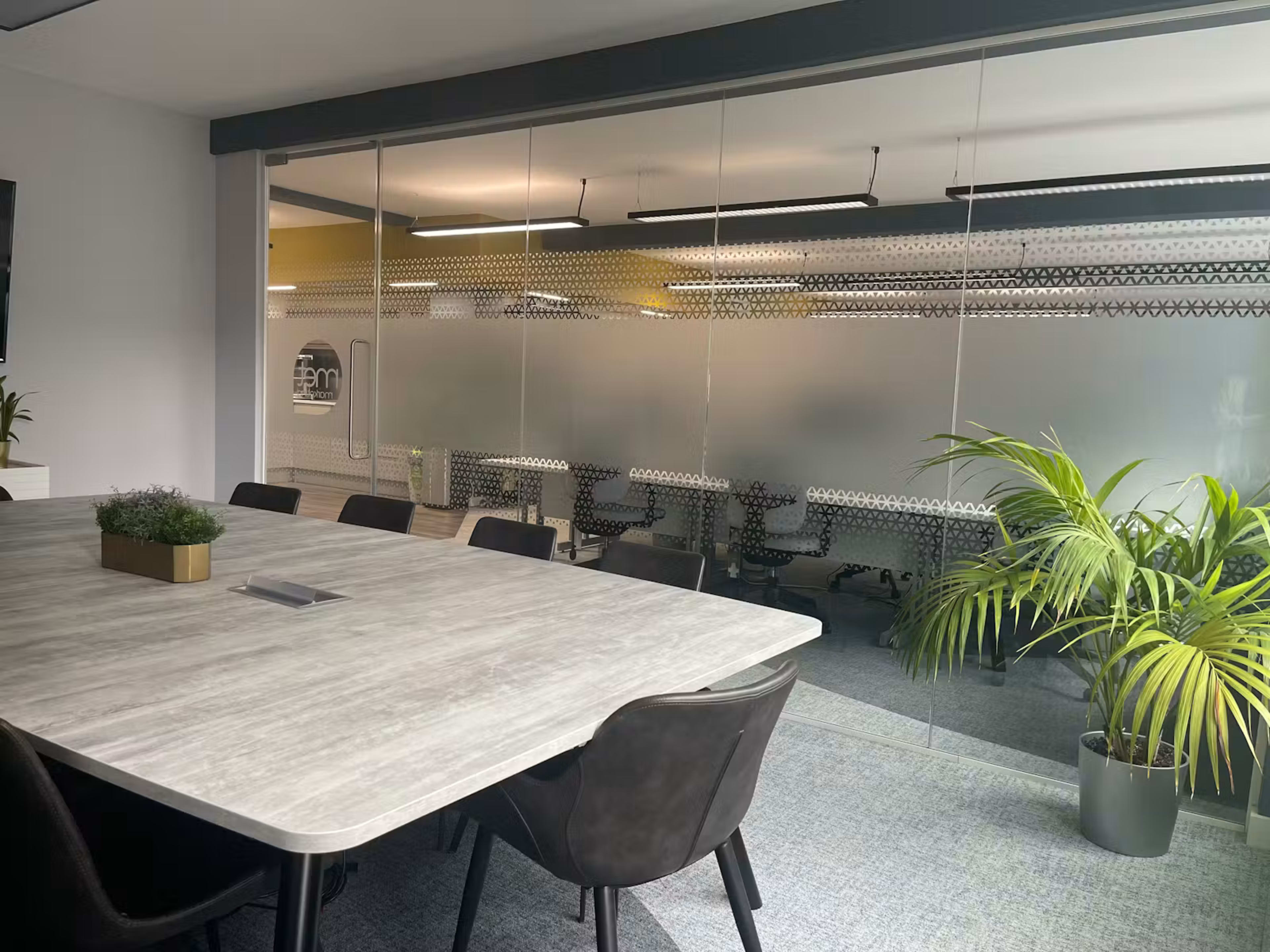 The image shows a modern office meeting room with a large table surrounded by black chairs, and a glass wall leading to another workspace with additional seating and decorative plants.
