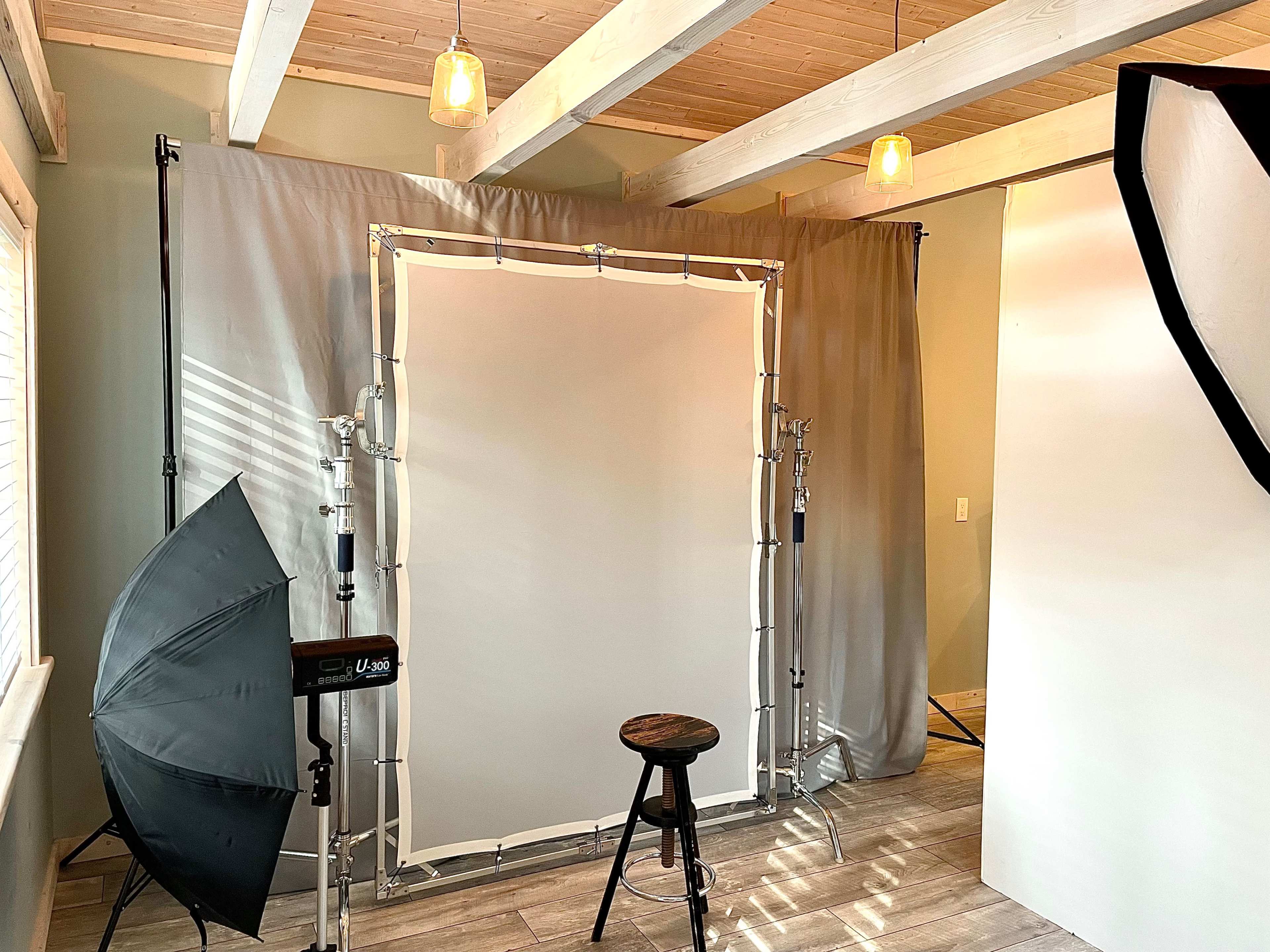 The image shows a photography setup featuring a backdrop with lighting equipment and a stool in a room with wooden beams and a light-colored floor.