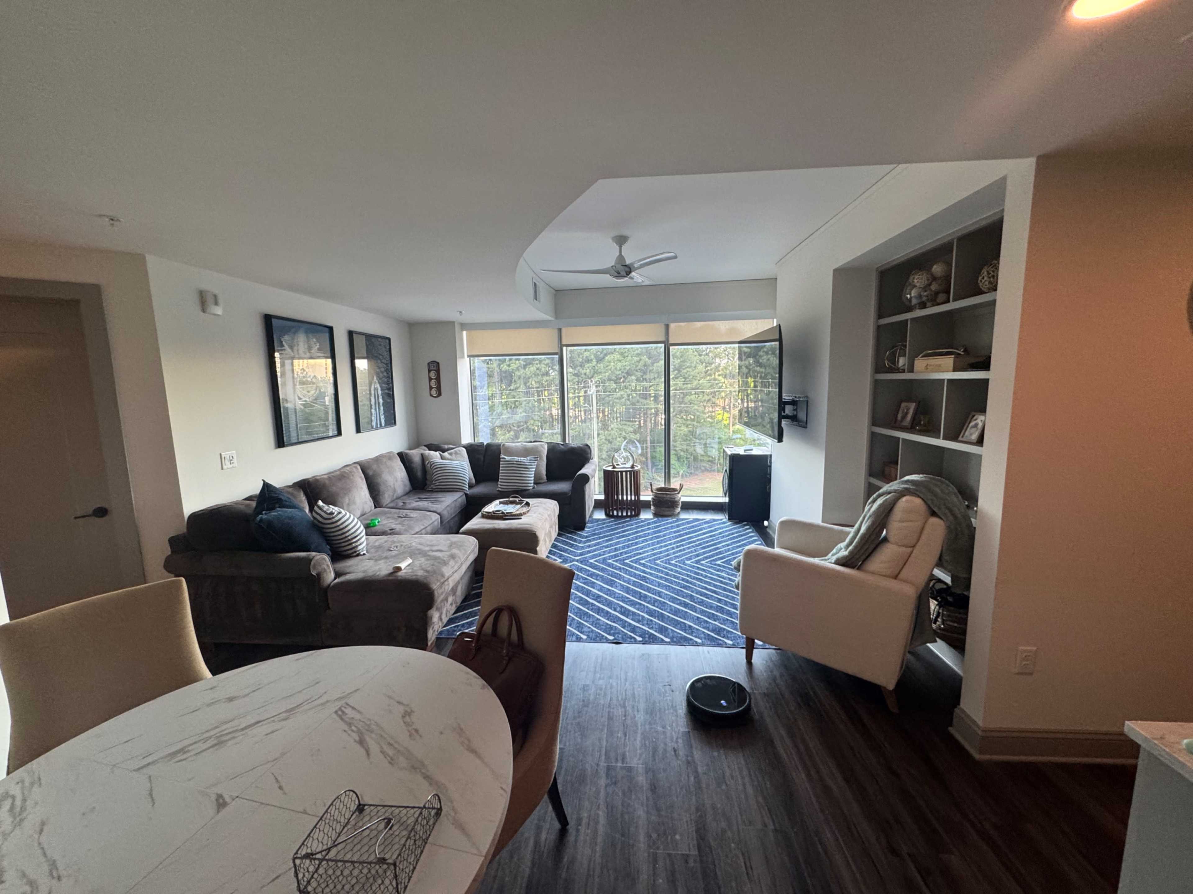 The image shows a modern living space that includes a seating area with a sofa and chairs, a dining table, and large windows overlooking a wooded area.