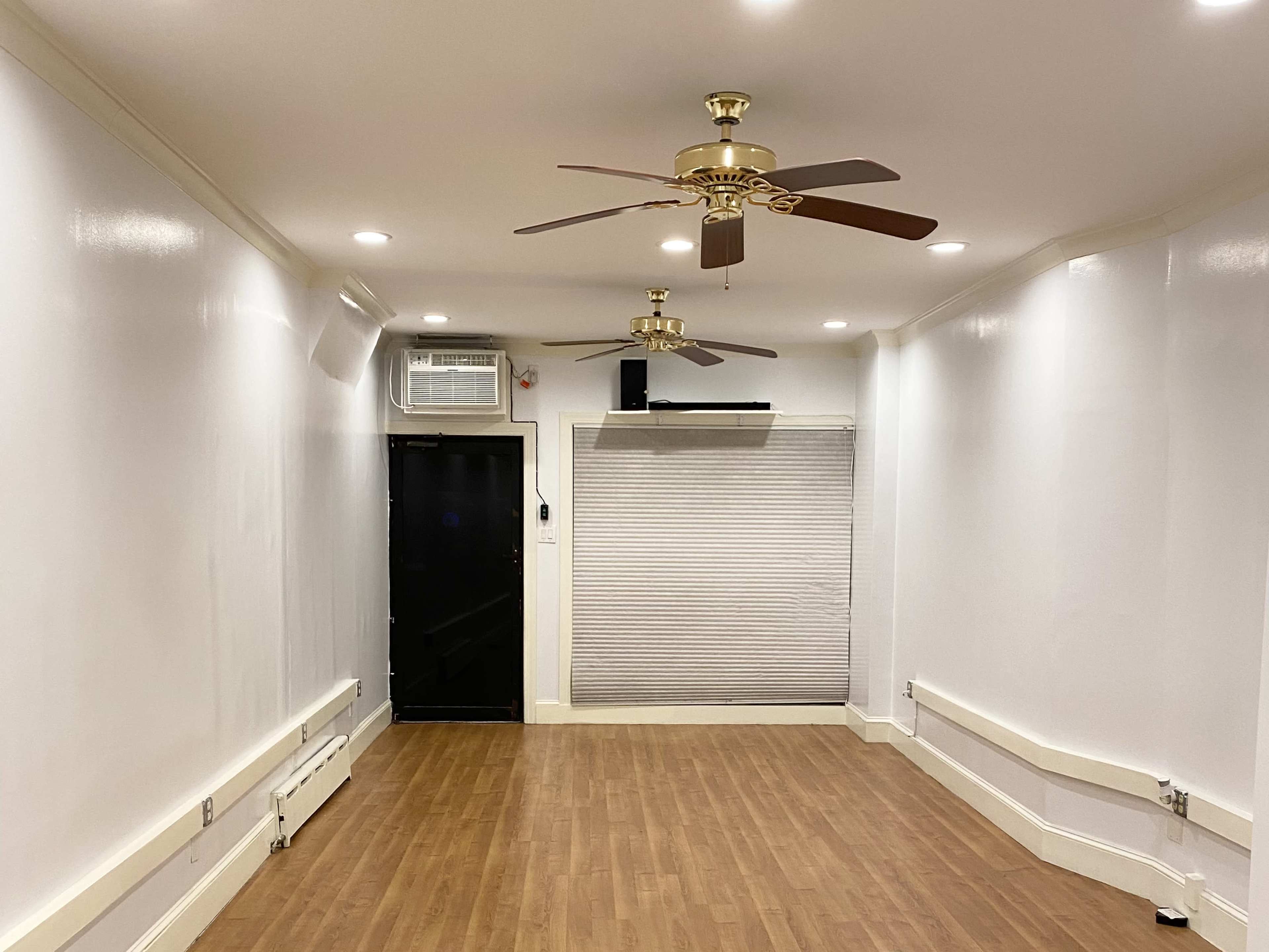 The image shows a room with wooden flooring, two ceiling fans, and a wall with a closed window covering.
