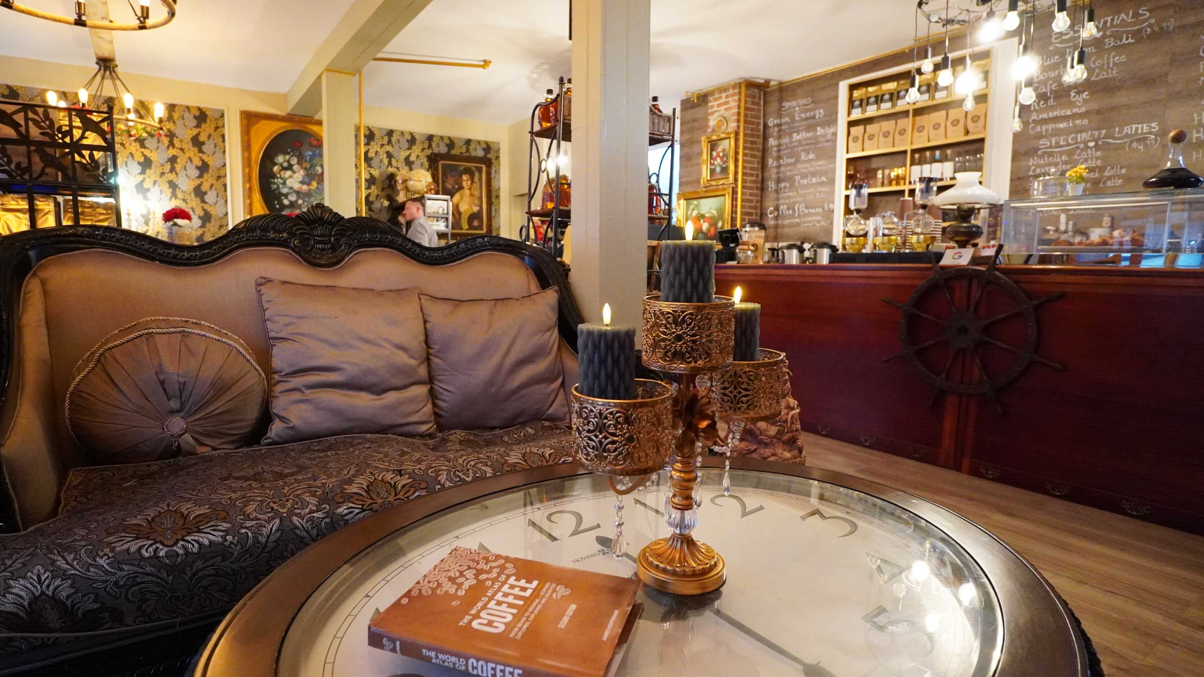 A cozy café interior featuring an ornate sofa, a table with a clock design, and a counter displaying an array of beverages and décor.