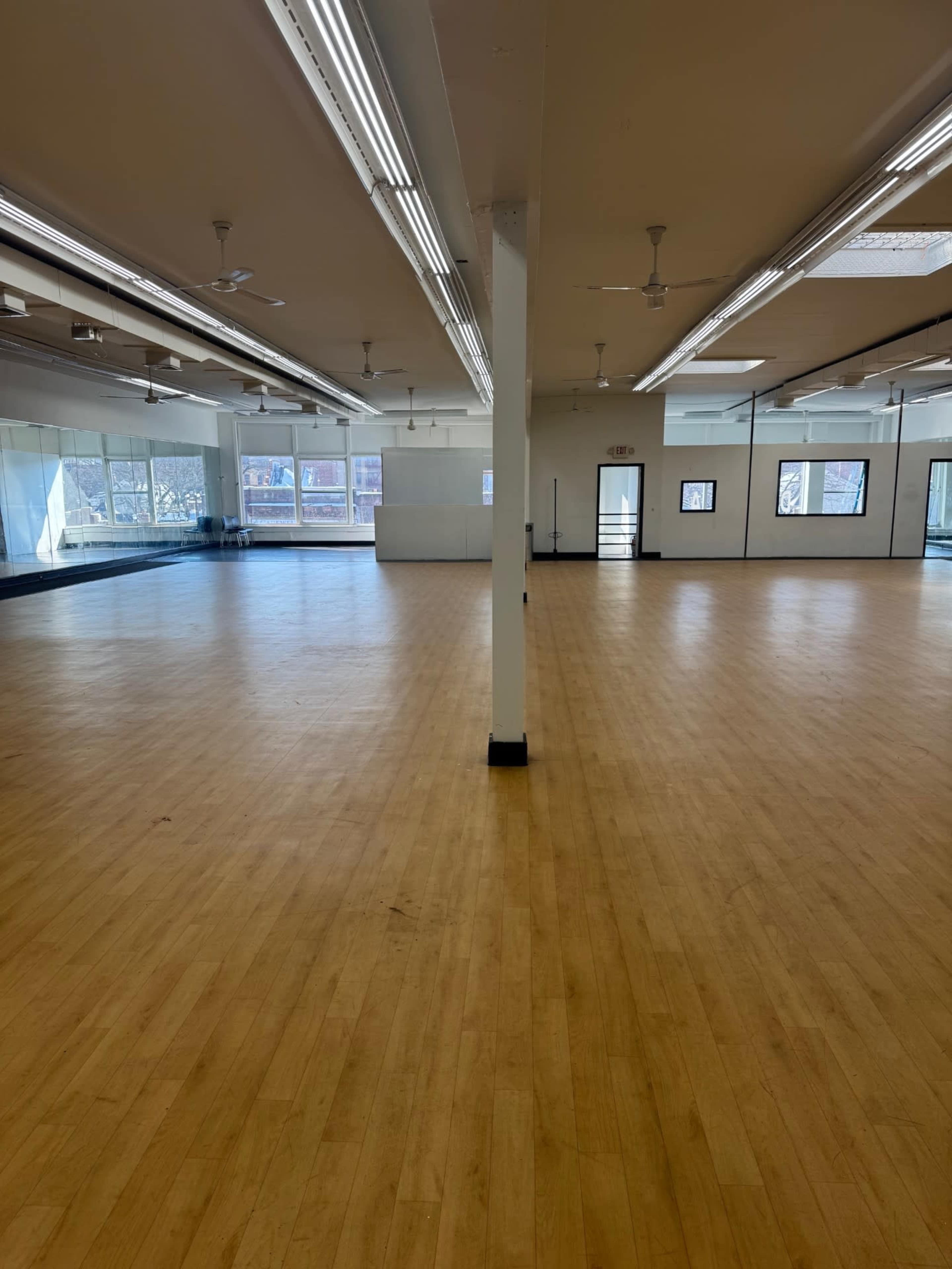 The image shows a spacious, empty indoor studio with wooden flooring, large windows, and bright overhead lights.