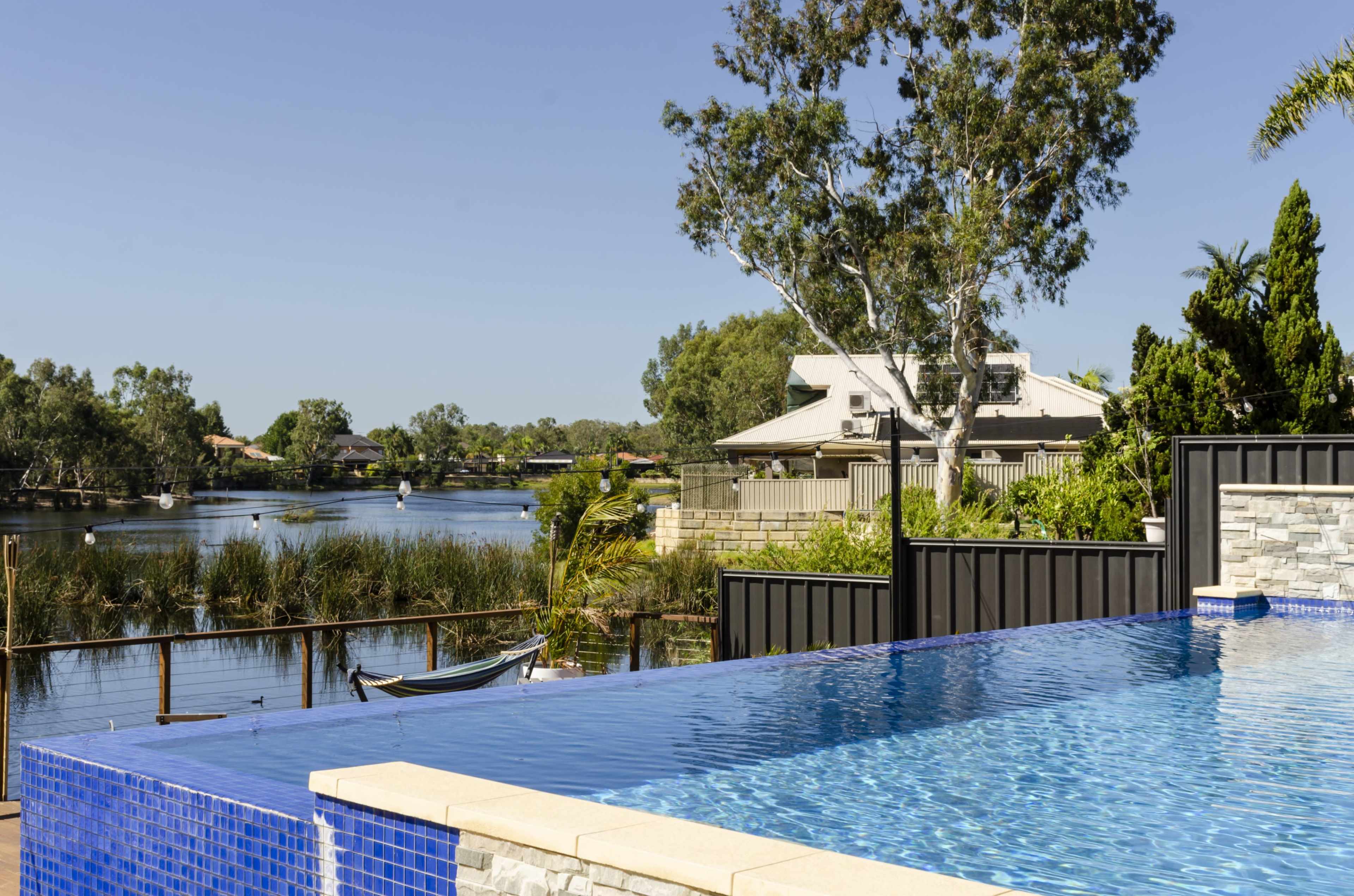 A modern infinity pool overlooks a calm river framed by lush greenery and residential buildings.