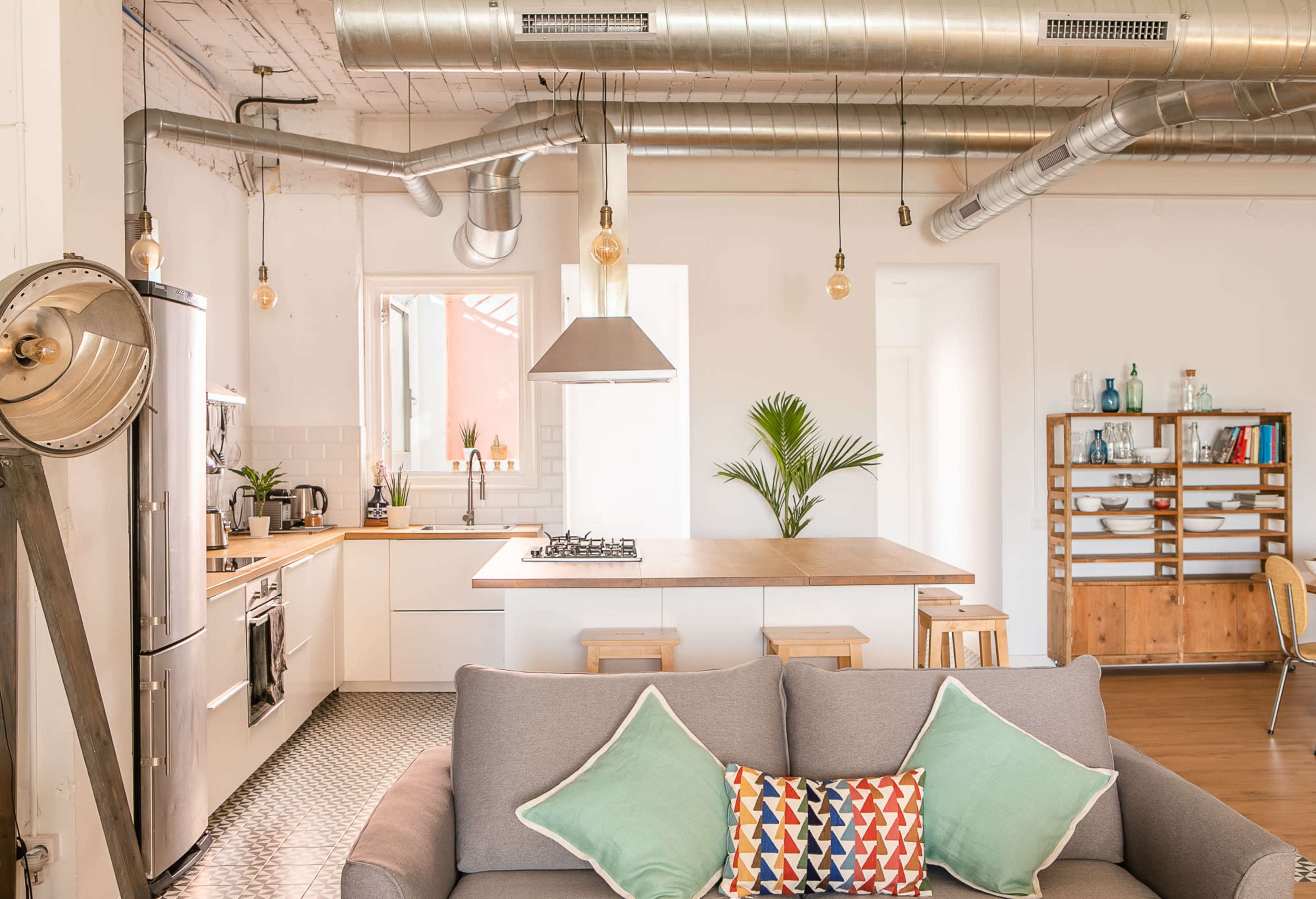 A modern kitchen and living area with exposed metal ductwork, a central island, and a sofa adorned with colorful cushions.