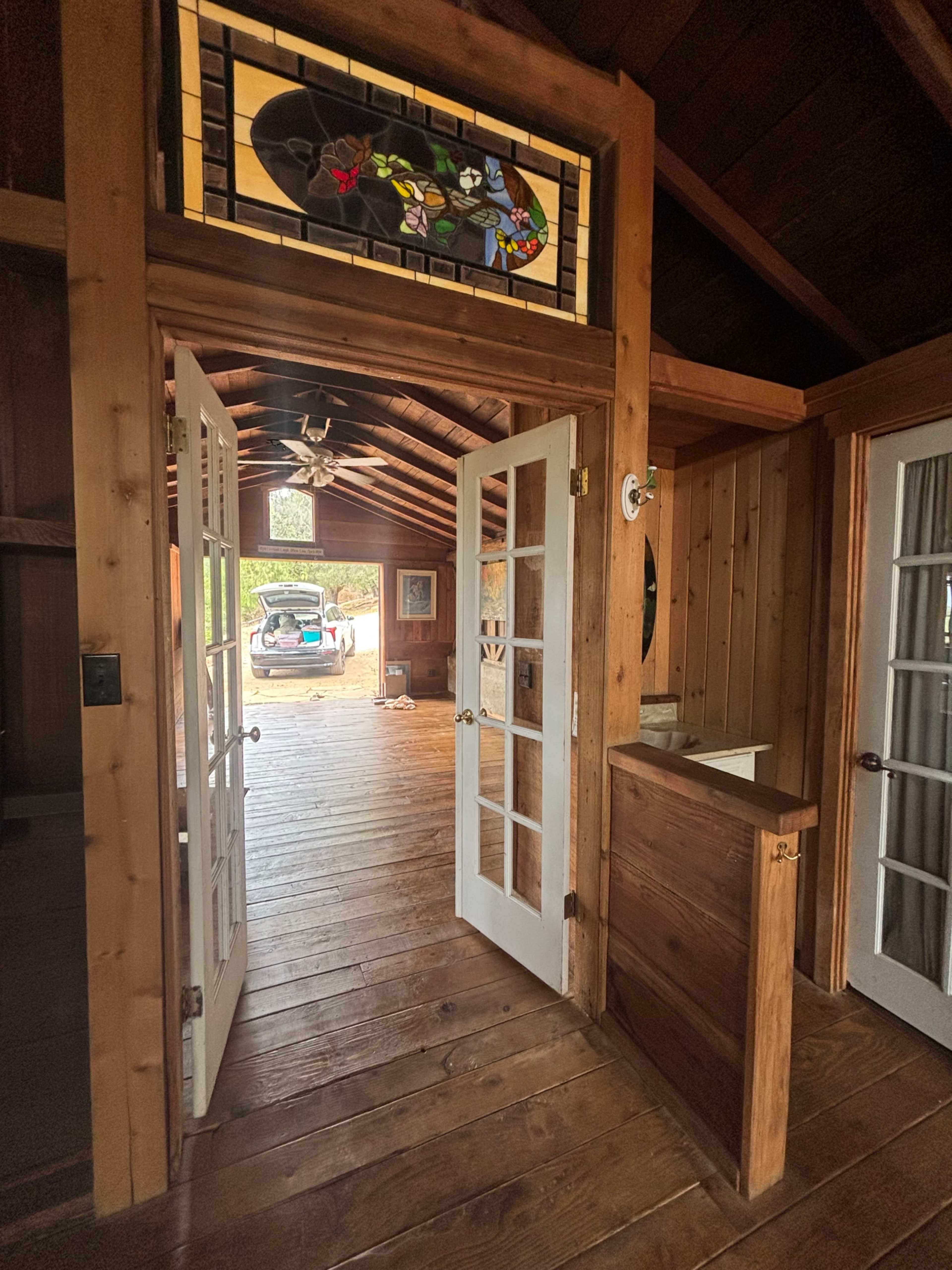 The image shows a wooden interior space with double doors leading outside, featuring a stained glass window above and a view of a vehicle parked outside.