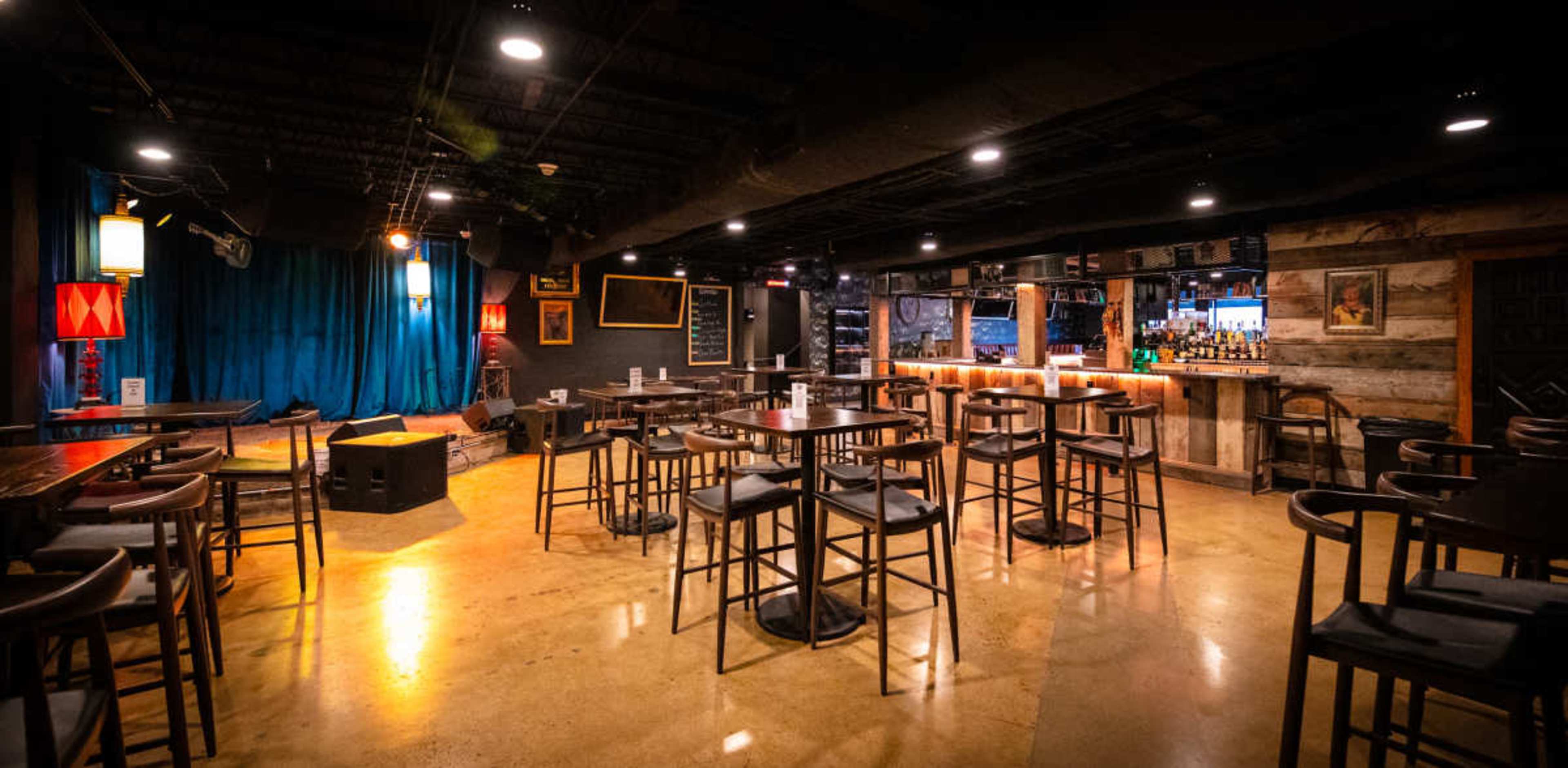 The image shows an empty bar area with wooden tables and stools, soft lighting, a stage with blue curtains, and a well-stocked bar in the back.