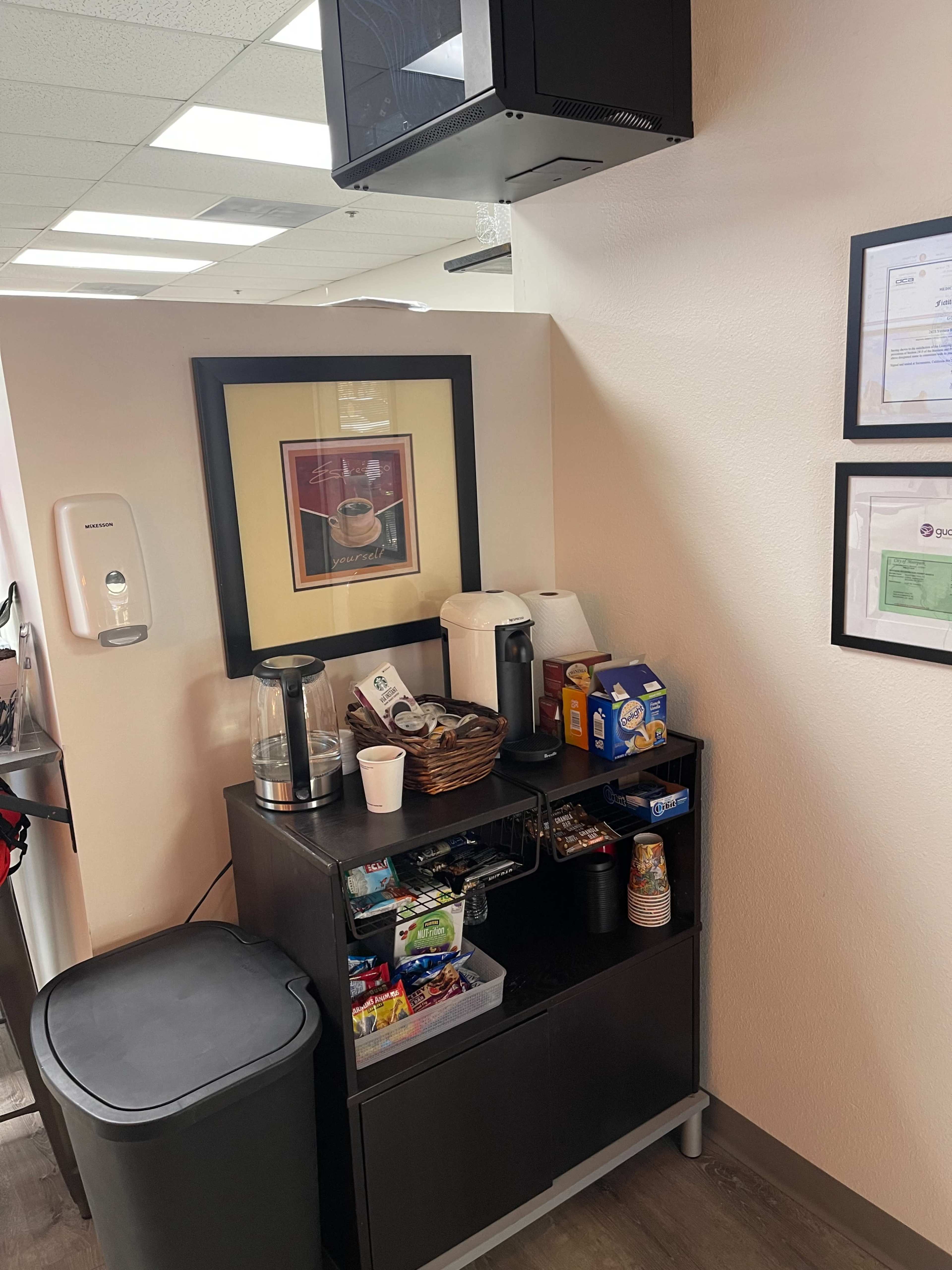 A small kitchenette area in an office, featuring a coffee maker, a basket of snacks, and various food items on a cabinet, beside a trash can and framed documents on the wall.