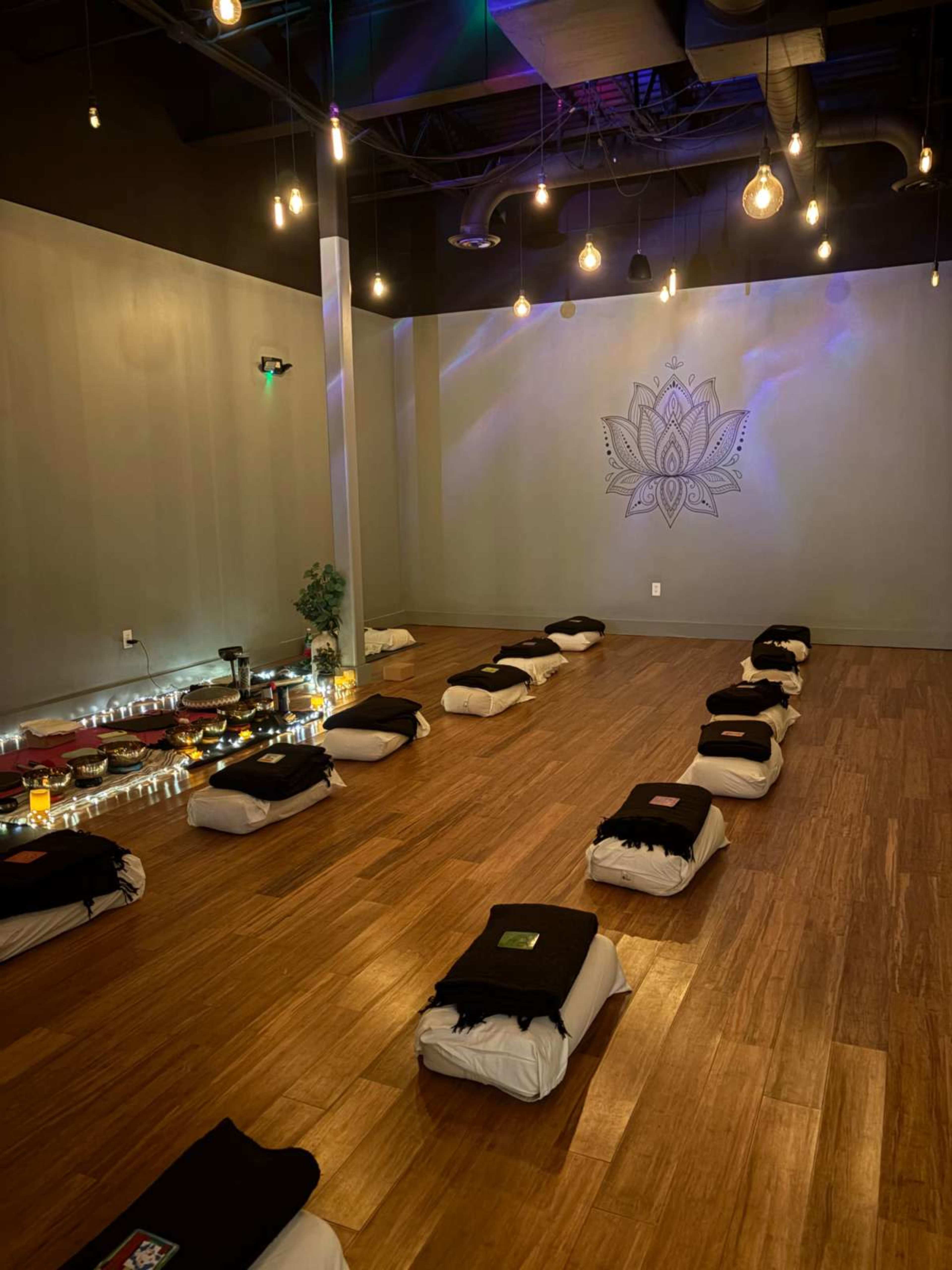 The image shows a dimly lit yoga studio with neatly arranged cushions on a wooden floor and a lotus mural on the wall.
