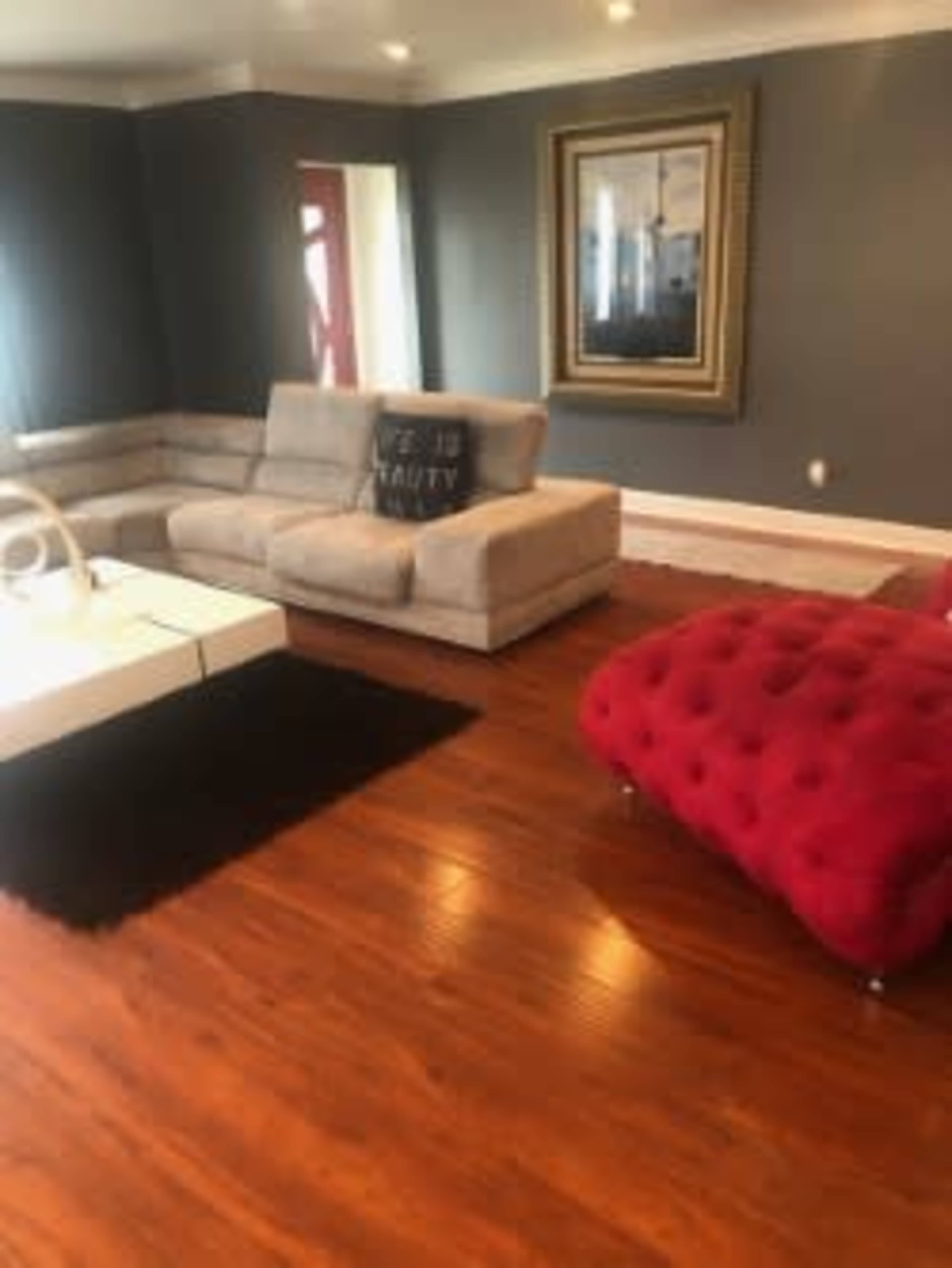 The image shows a modern living room with a gray wall, a light-colored sectional sofa, a red tufted ottoman, and a rectangular coffee table on a dark rug.