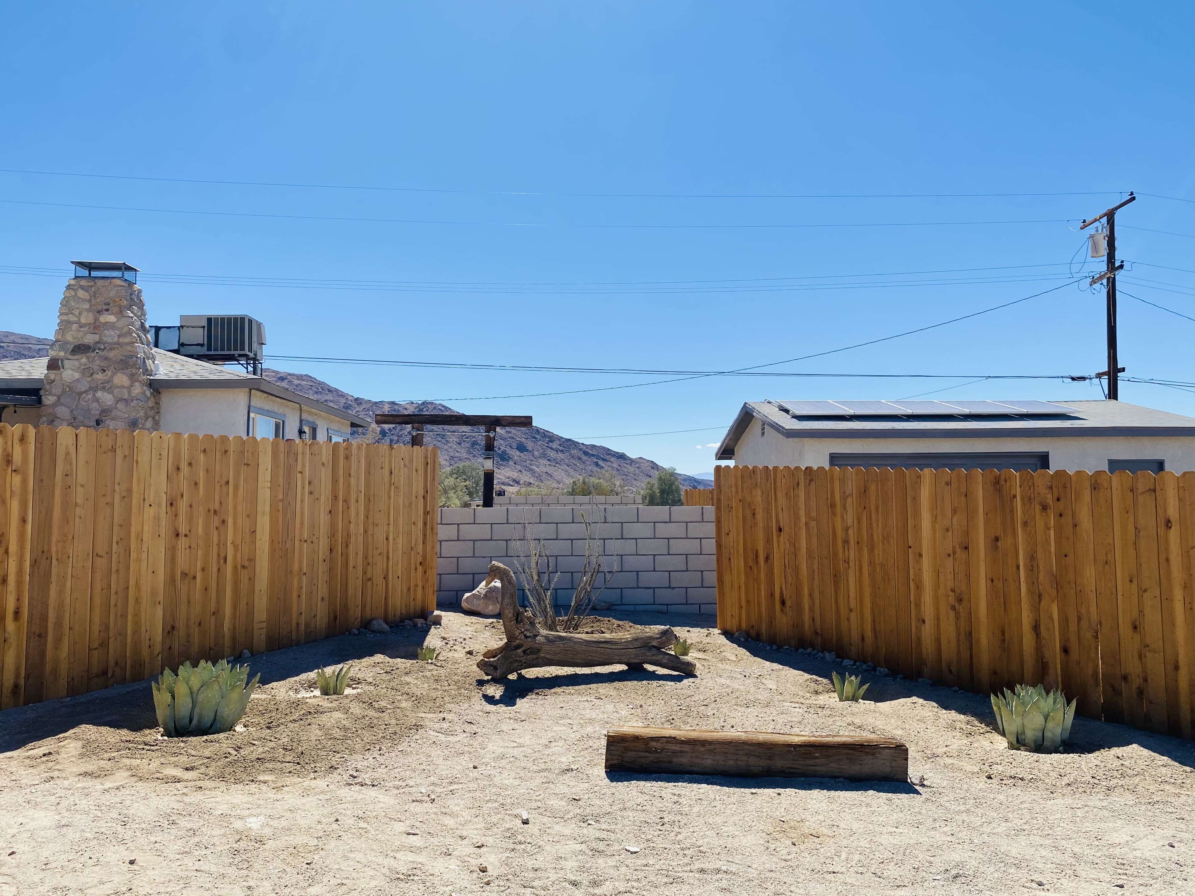 A cleared, sandy yard surrounded by wooden fencing, with a log and a piece of driftwood placed on the ground and a backdrop of distant hills.