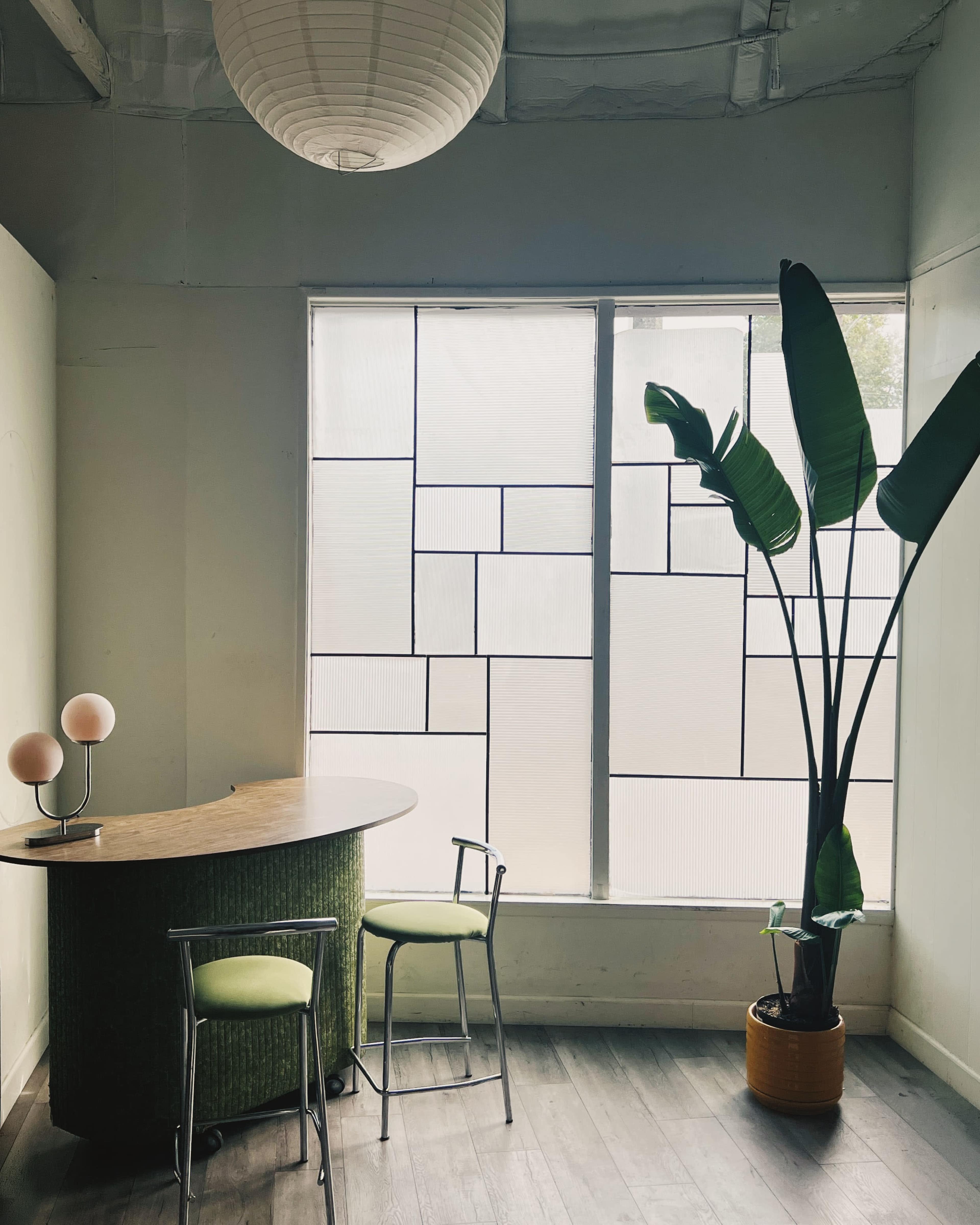 The image shows a modern interior with a round table and two chairs beside a large window, which features a geometric pattern, and a tall potted plant in the corner.