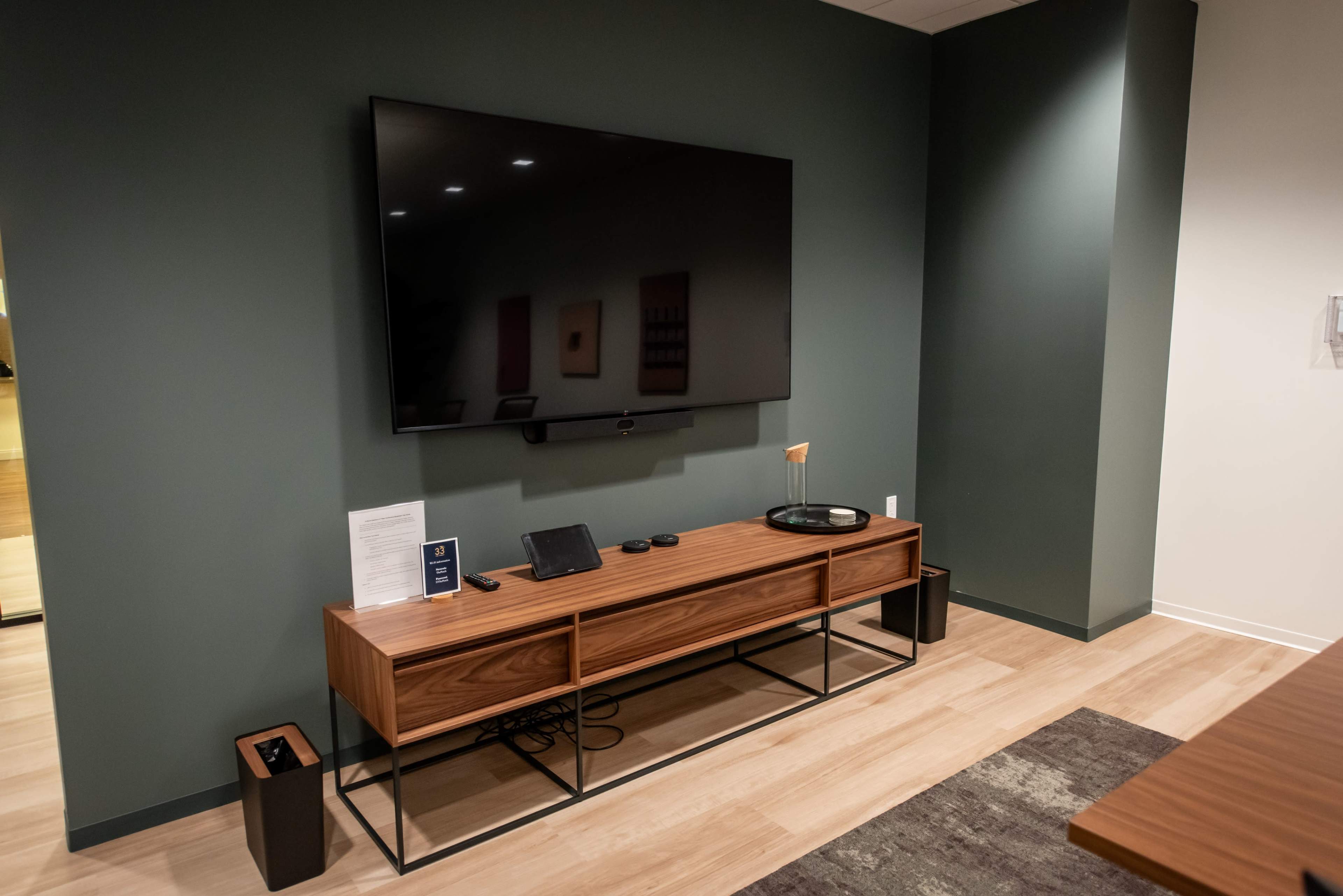 The image shows a modern living room setup featuring a large flat-screen TV mounted on a green wall above a sleek wooden media console, which contains decorative items and electronic devices.