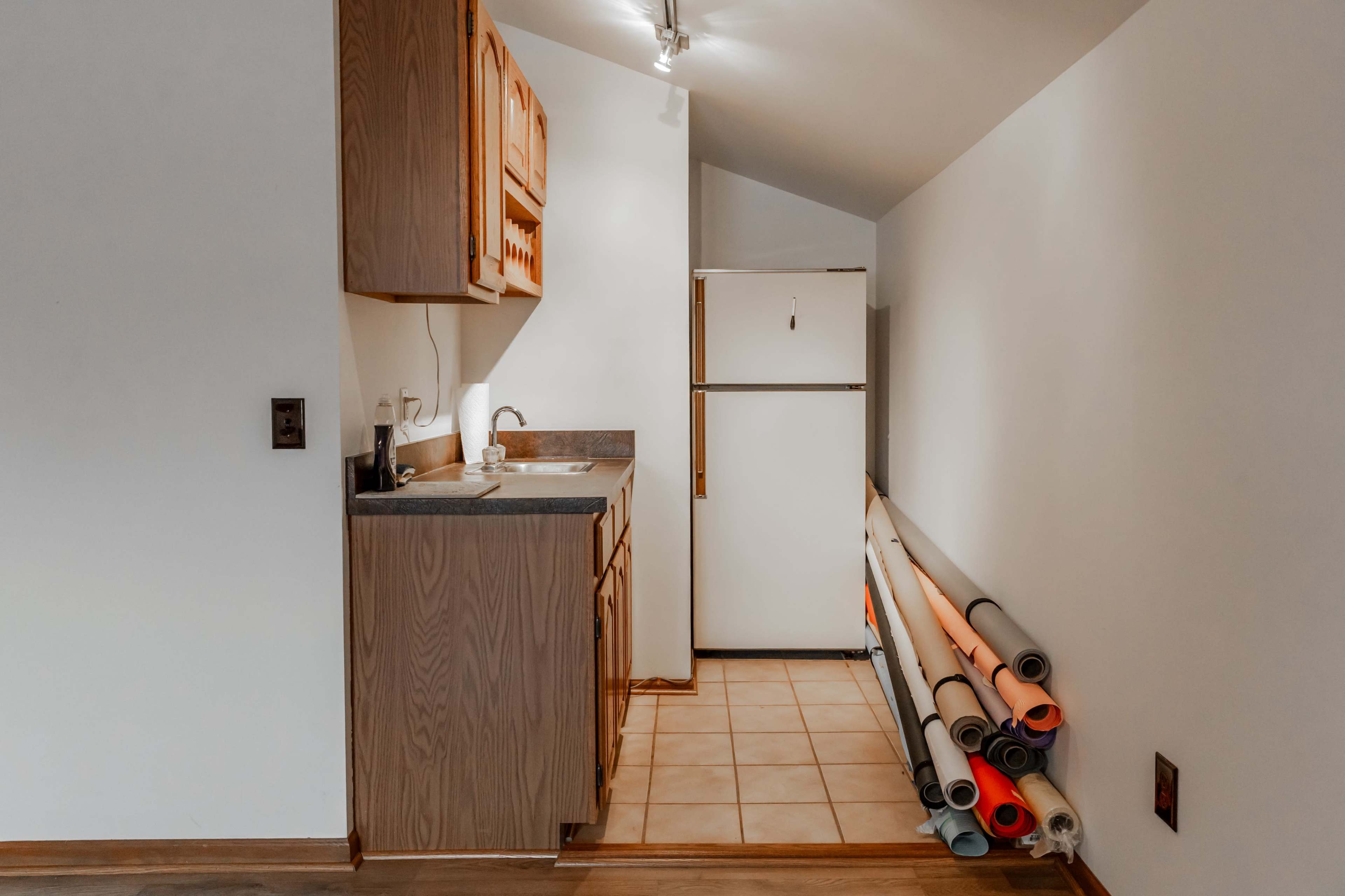 A narrow kitchen space with wooden cabinets, a sink, a refrigerator, and rolled-up mats stacked against the wall.