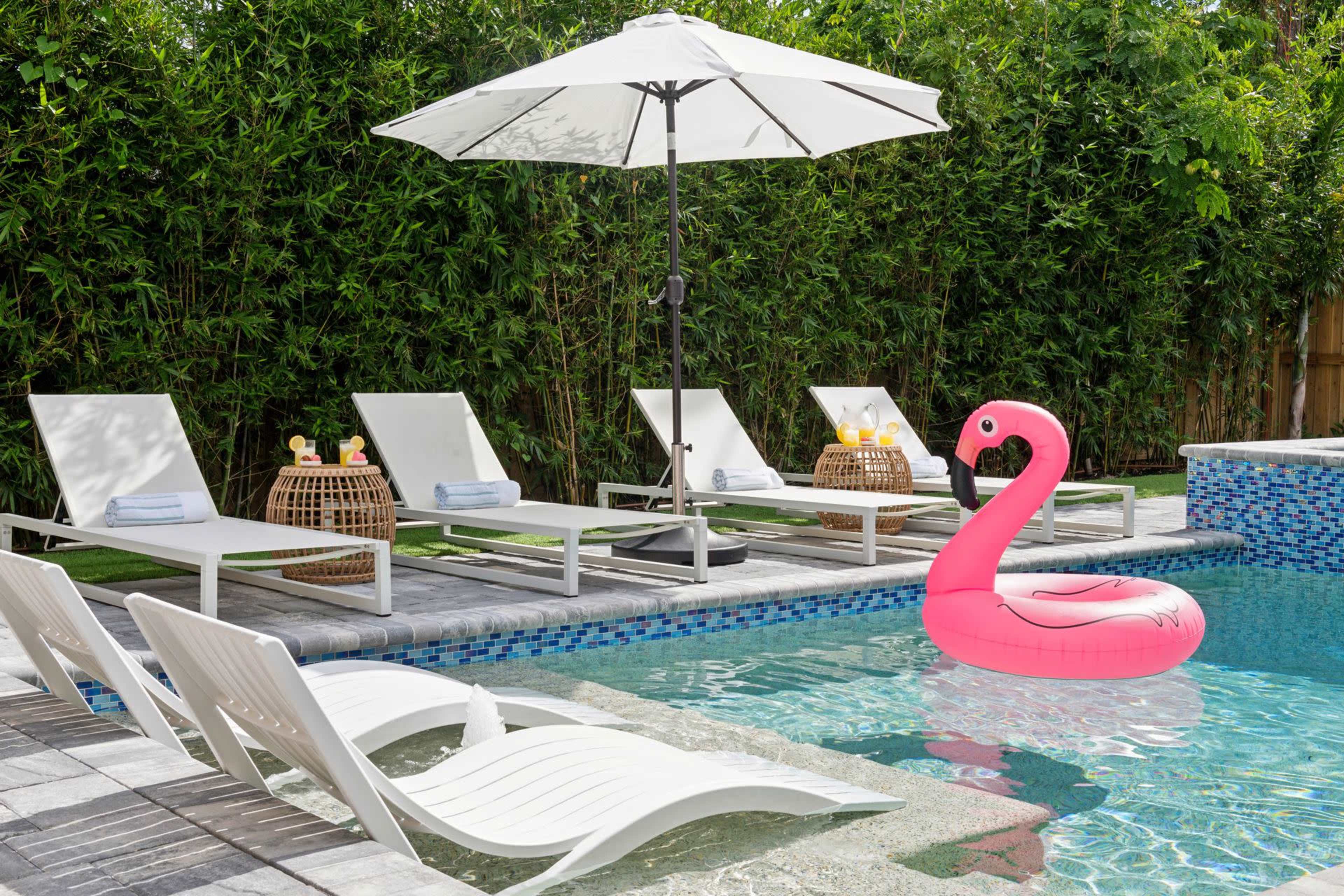 The scene features a swimming pool with a pink flamingo float, surrounded by white lounge chairs, an umbrella, and decorative baskets.