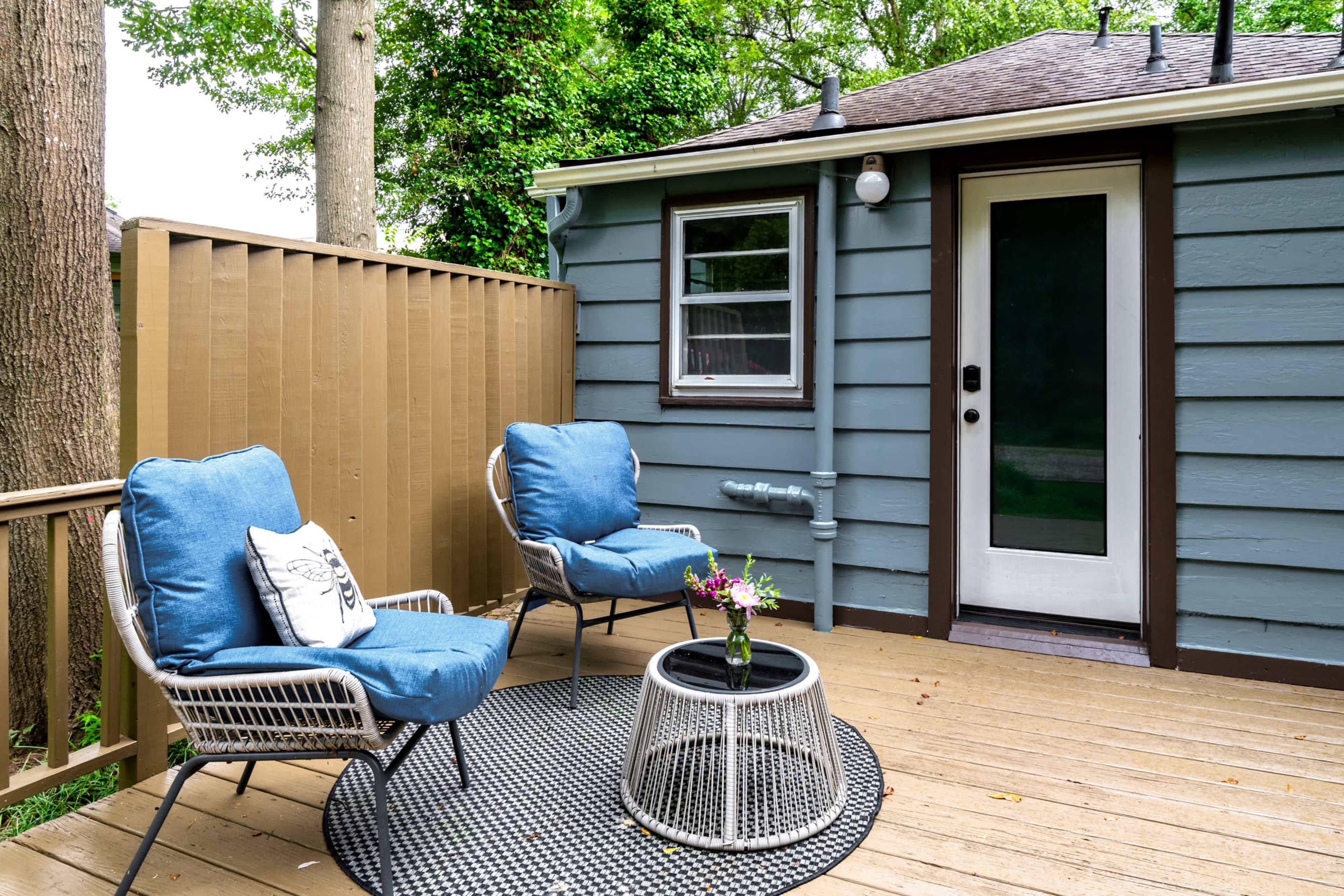 A small deck features two blue armchairs and a round table with a flower vase, adjacent to a light gray house with a door and window.