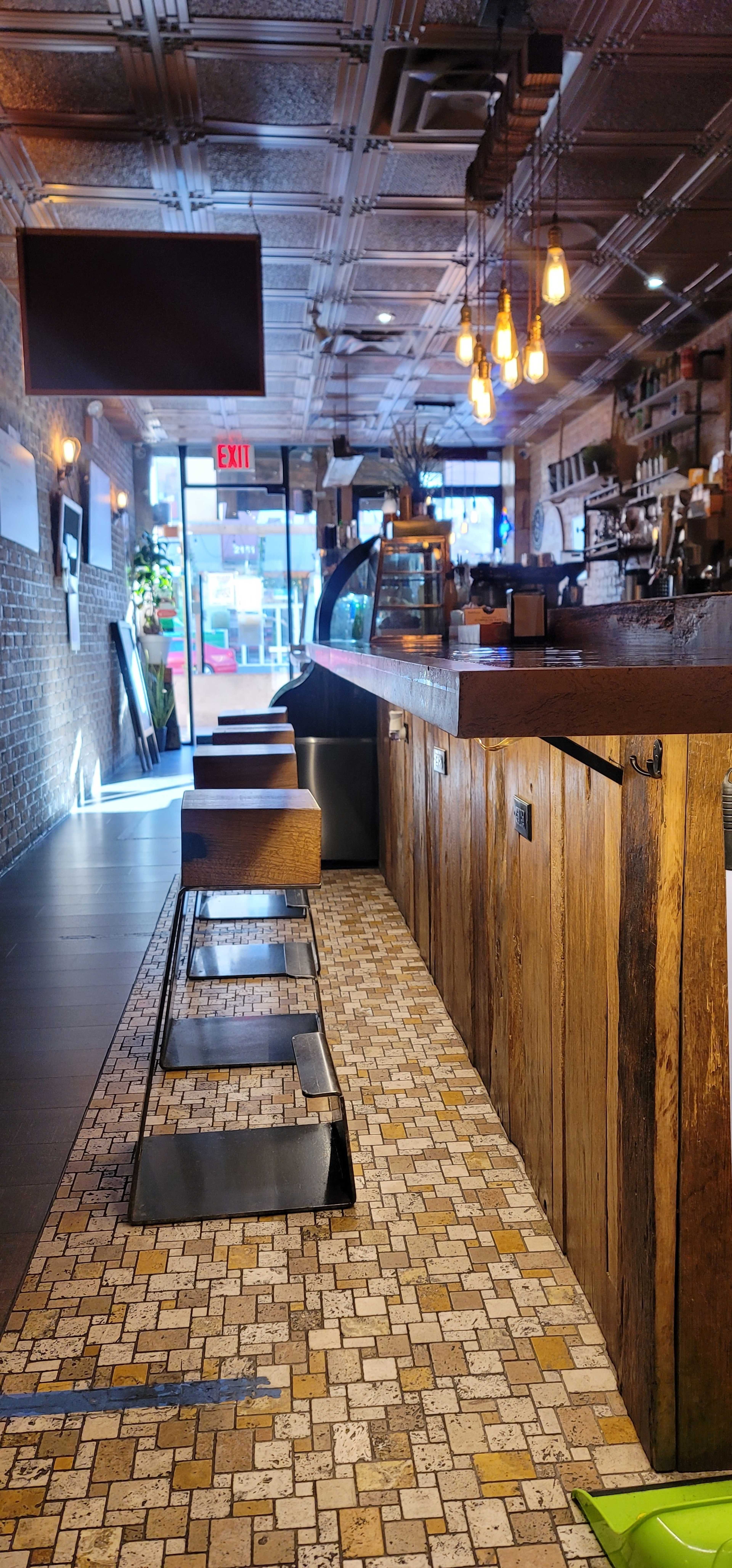A coffee shop interior with wooden bar seating, tiled flooring, and large windows allowing natural light to enter.