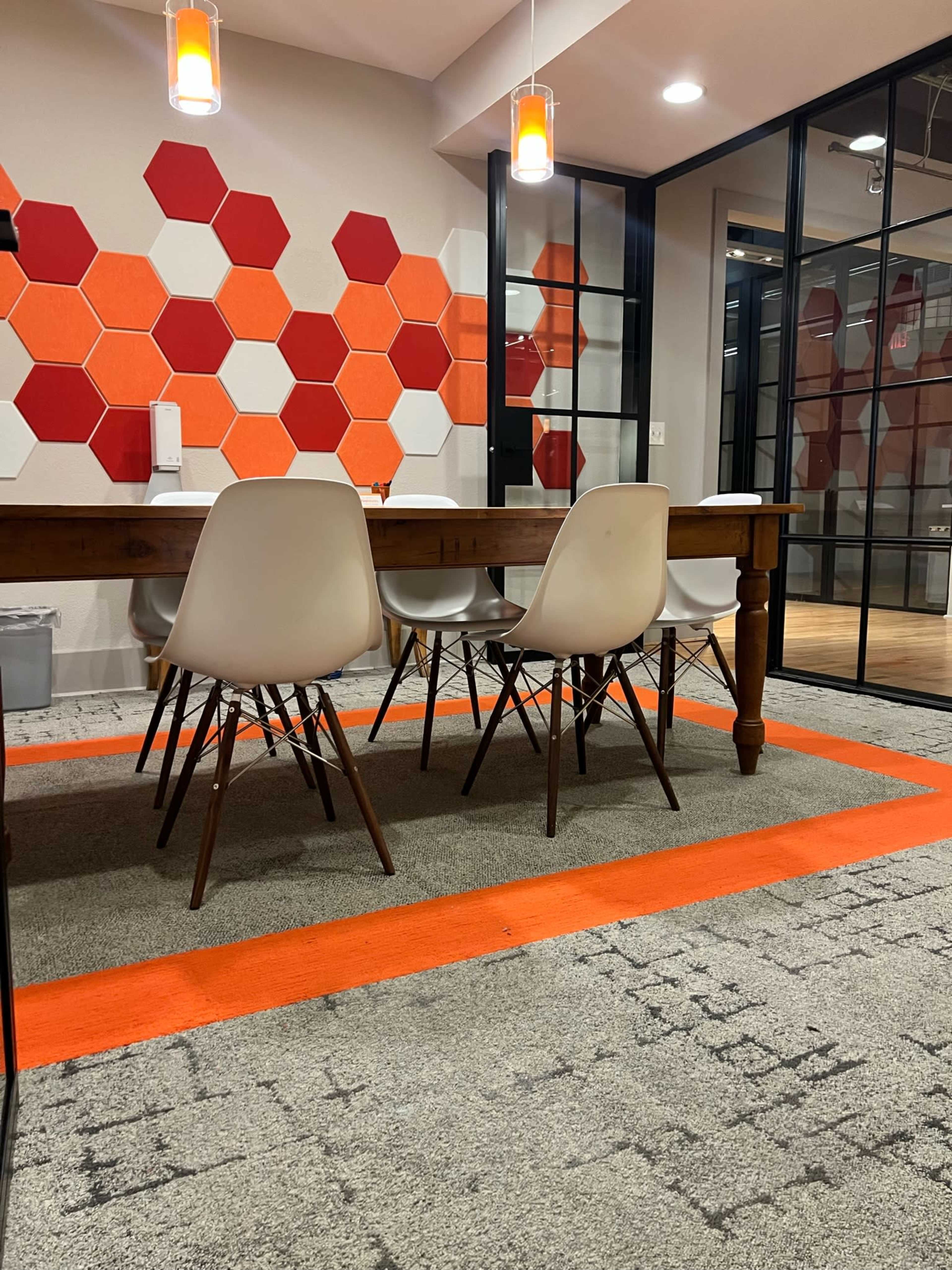 The image shows a conference room with a wooden table surrounded by white chairs and a hexagonal orange and red accent wall.