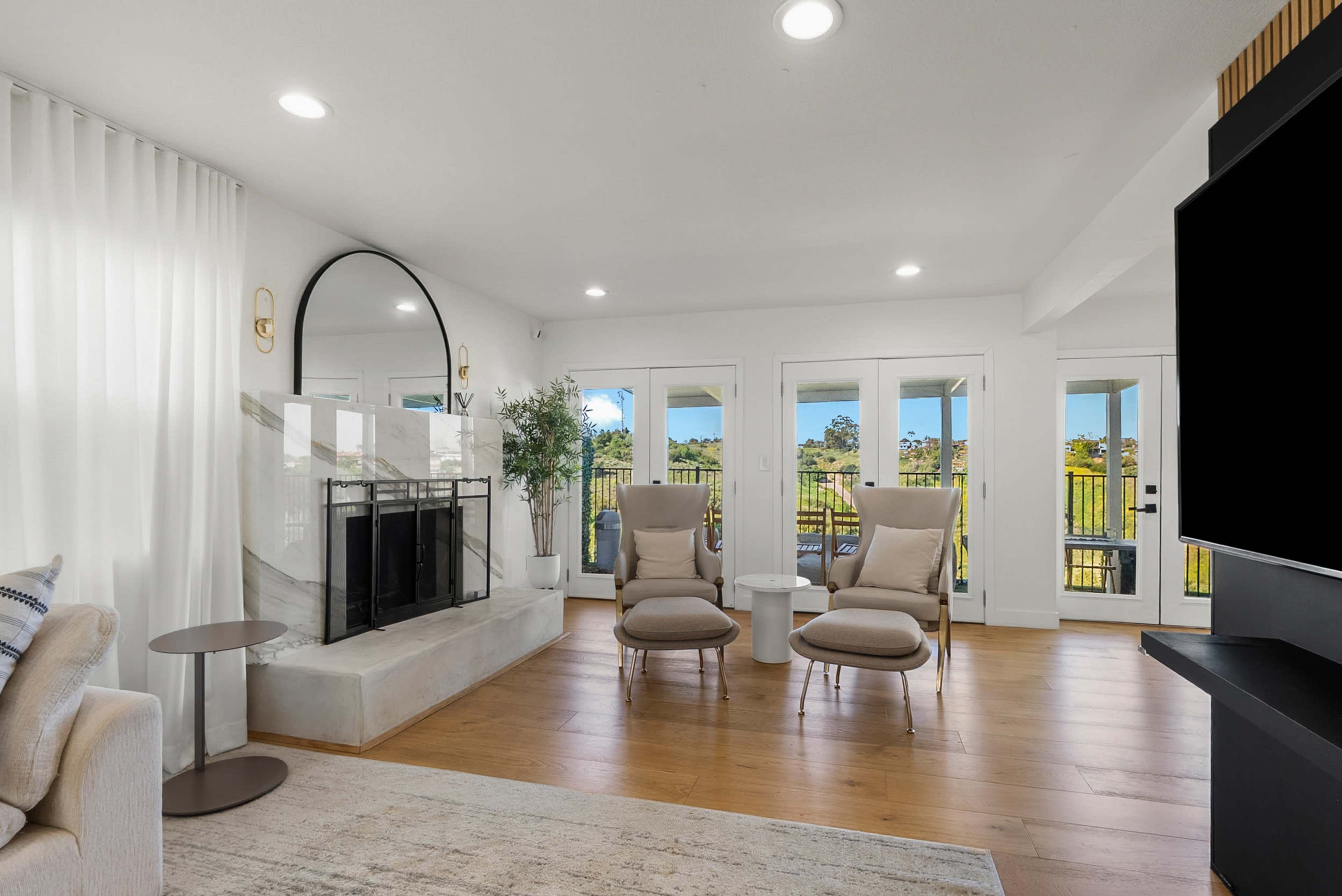 The living room features two beige armchairs facing a marble fireplace, with large windows allowing a view of the outdoors.