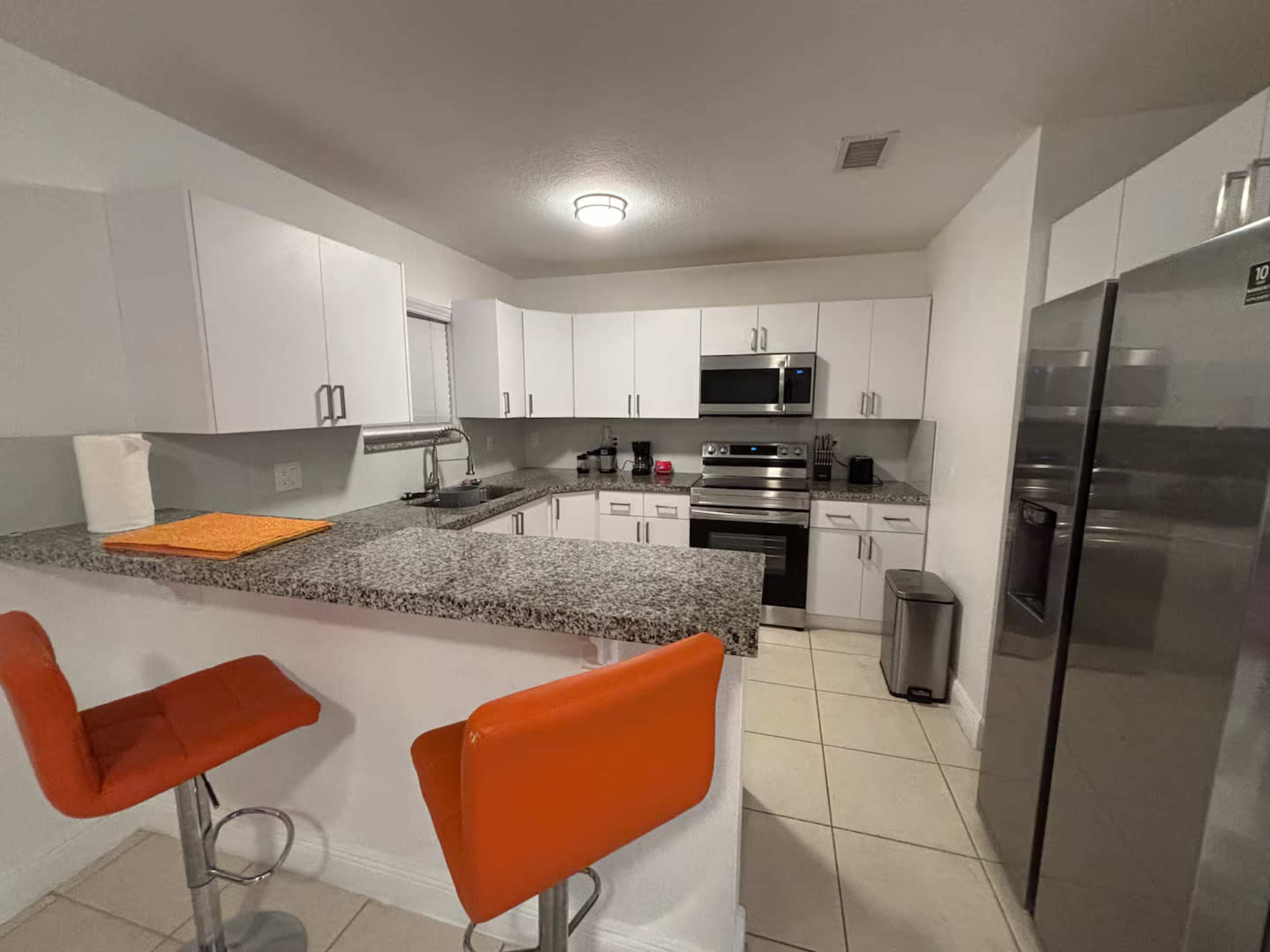The image shows a modern kitchen with white cabinetry, stainless steel appliances, a granite countertop, and two orange bar stools at the island.