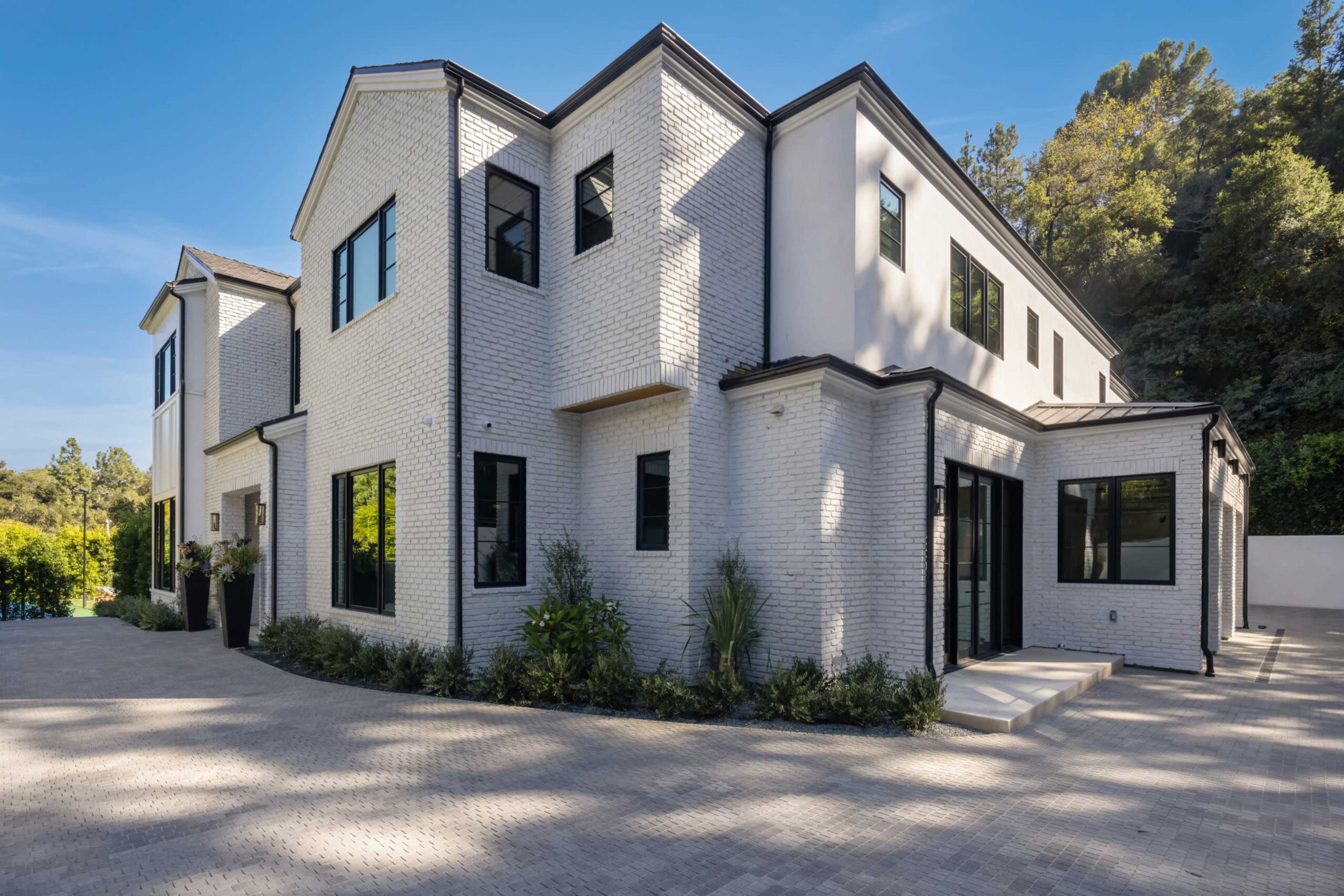 The image shows a modern, two-story white brick house surrounded by landscaped greenery and a paved driveway.
