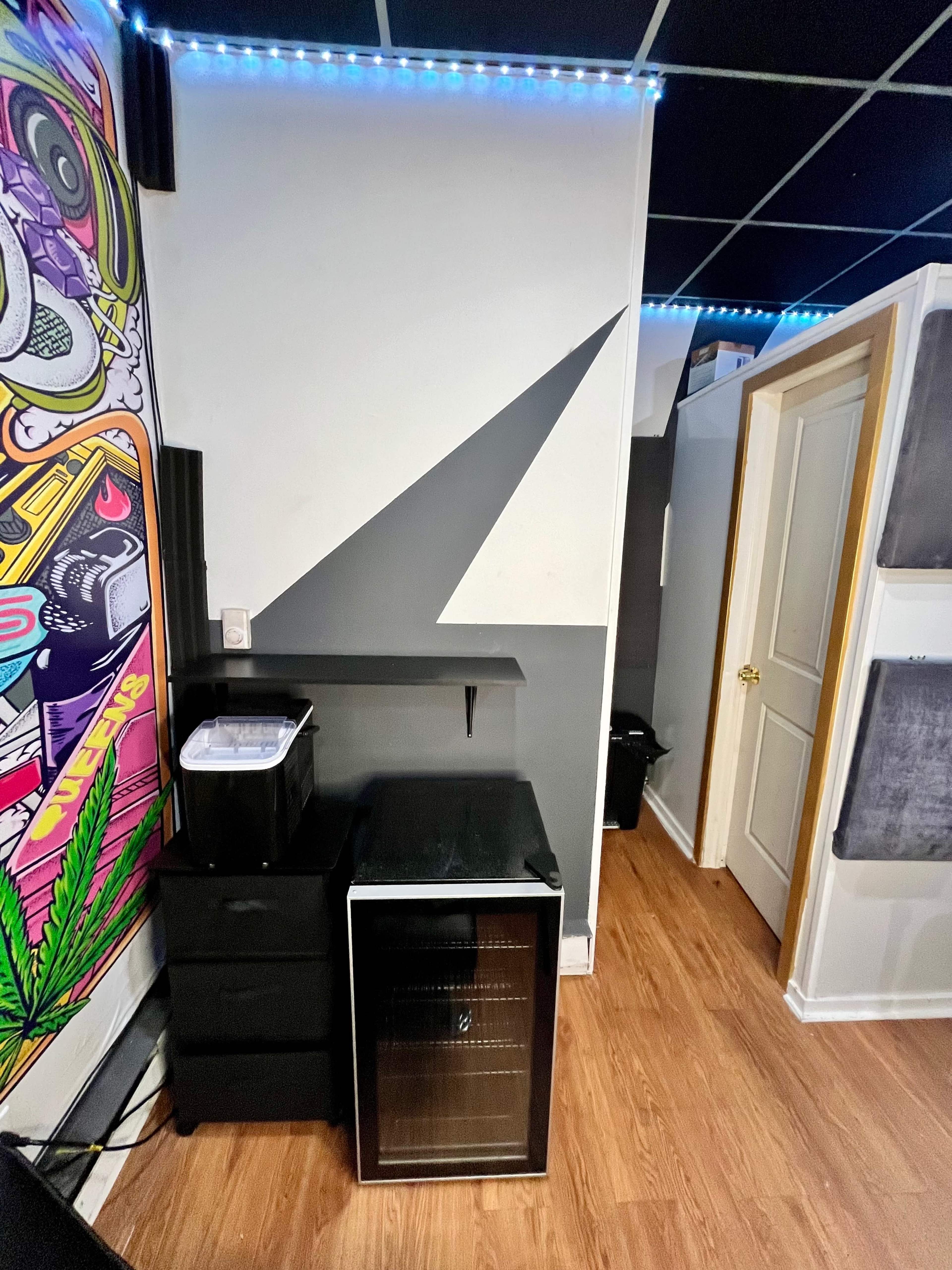 The image shows a wall with a colorful mural, a black shelf with a printer, and a small refrigerator positioned near a doorway with a white frame.