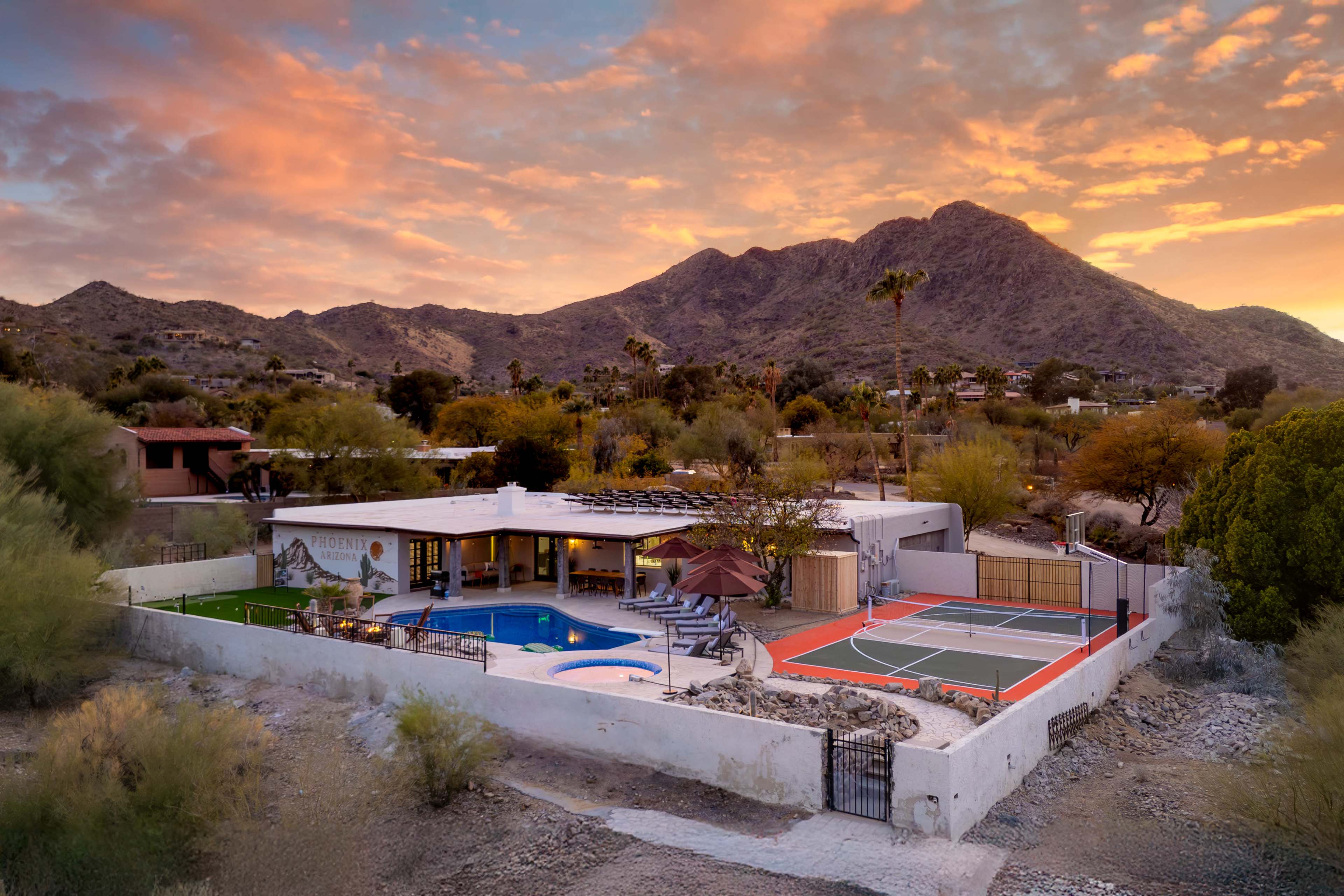 The image shows a modern home surrounded by desert landscaping, featuring a swimming pool, sports courts, and mountains in the background under a colorful sky.