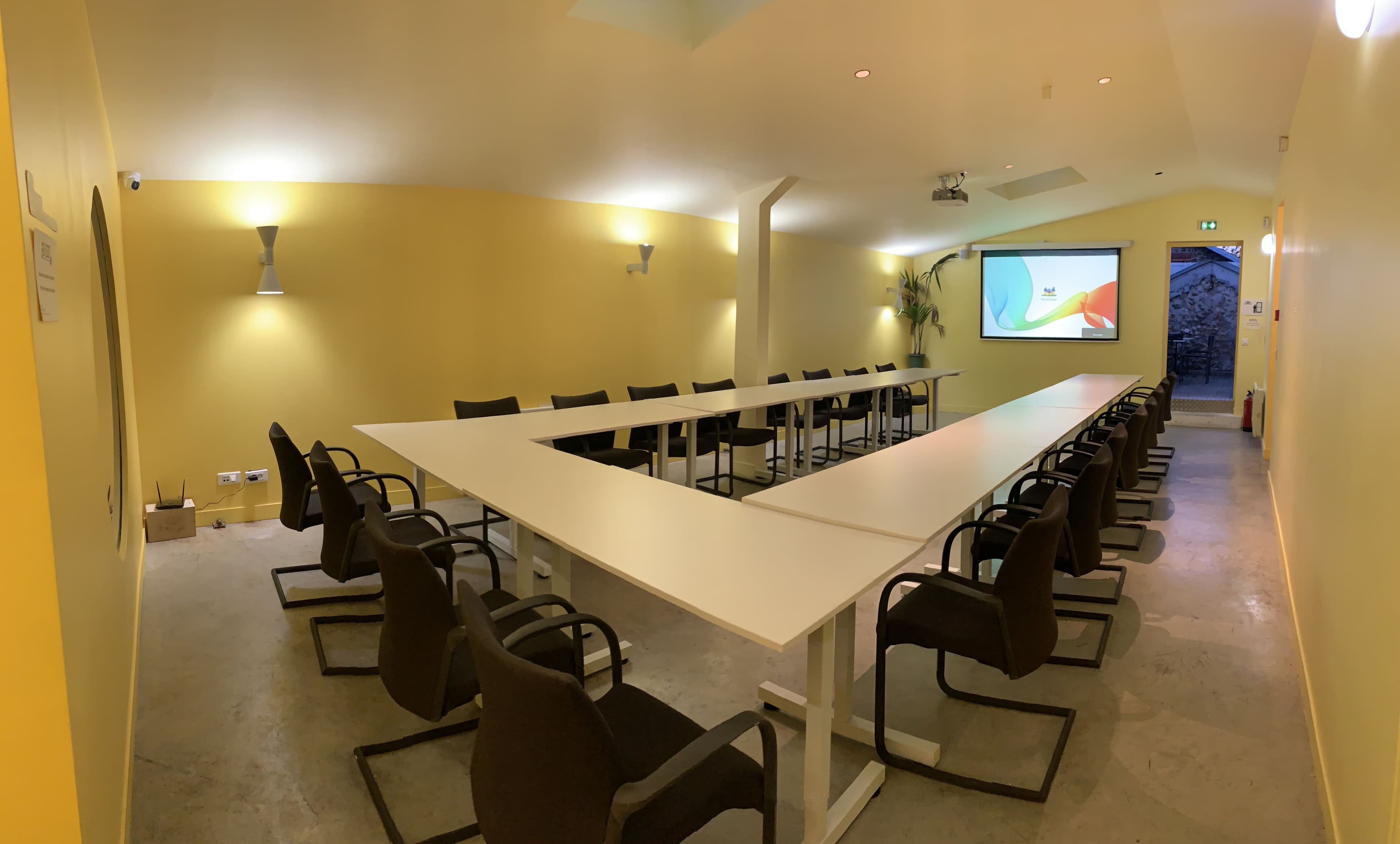 A modern conference room with a long, white table, black chairs, and a projector screen on a yellow wall.