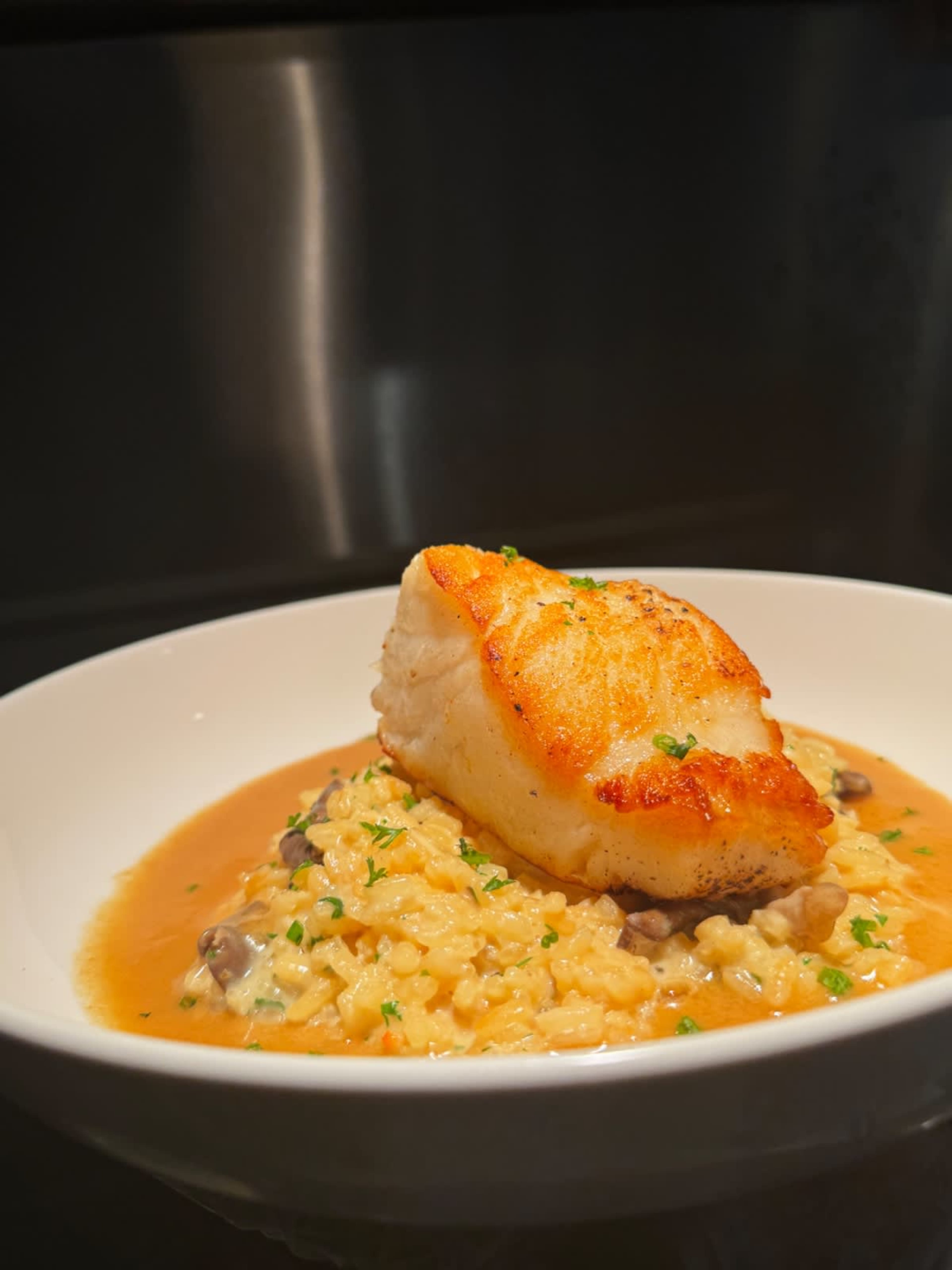 A piece of seared fish is placed on a bed of creamy risotto garnished with parsley in a white bowl.