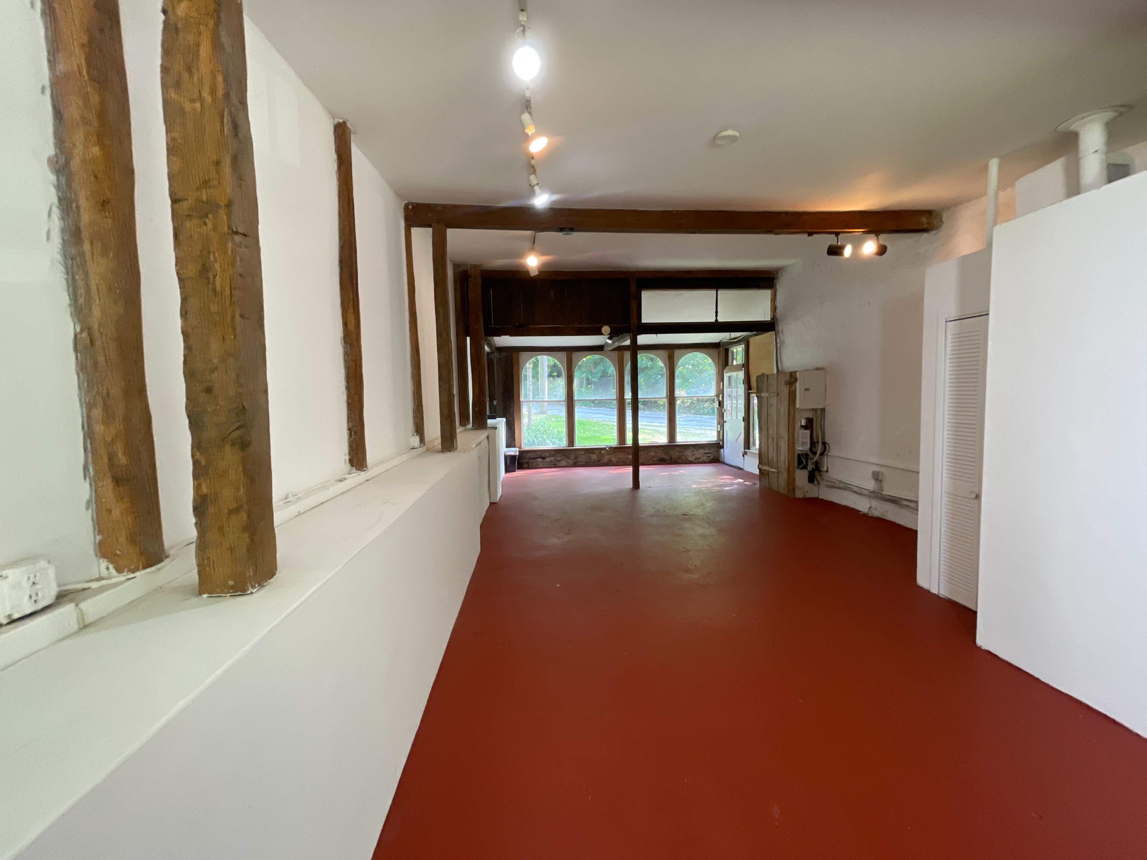 An empty interior space with red flooring, exposed wooden beams, and large windows at the far end.