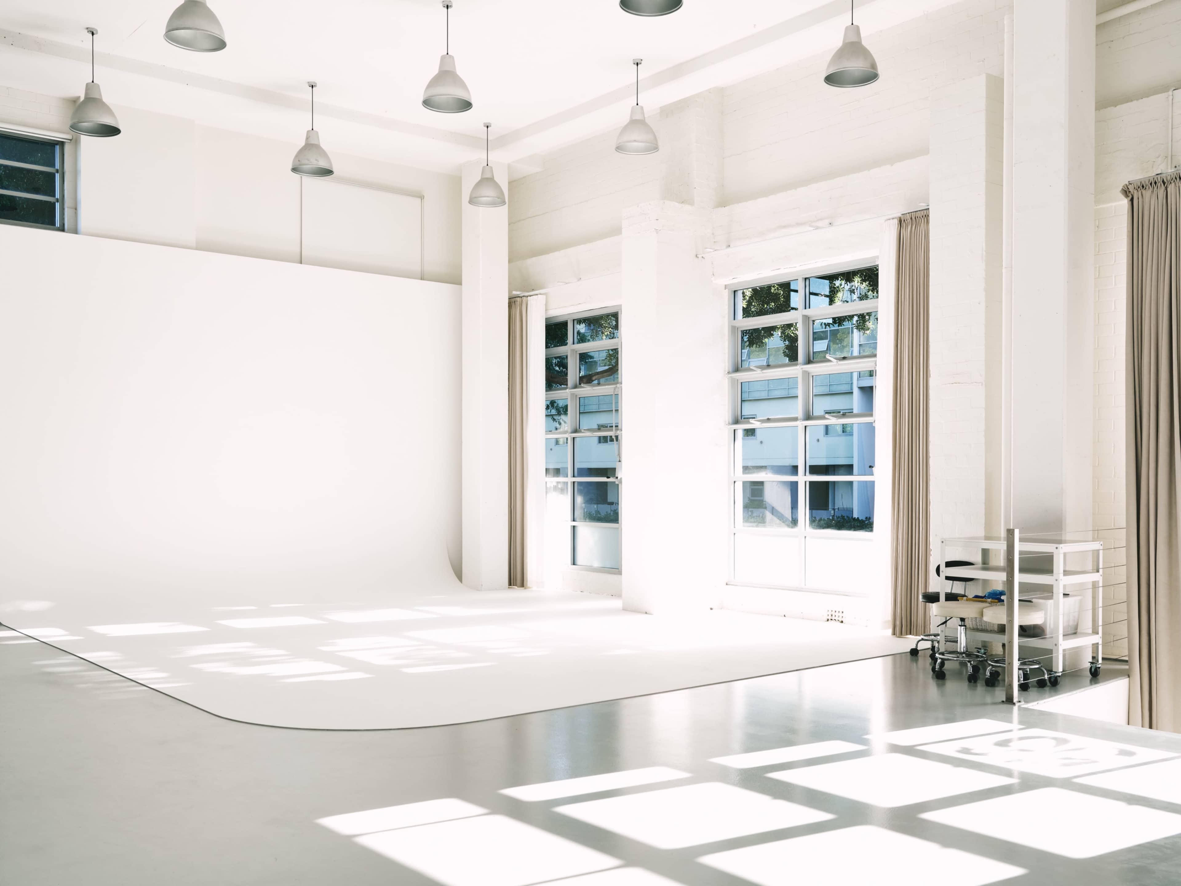 The image shows a spacious, well-lit photography studio with a white backdrop, large windows, and soft natural light casting shadows on the floor.