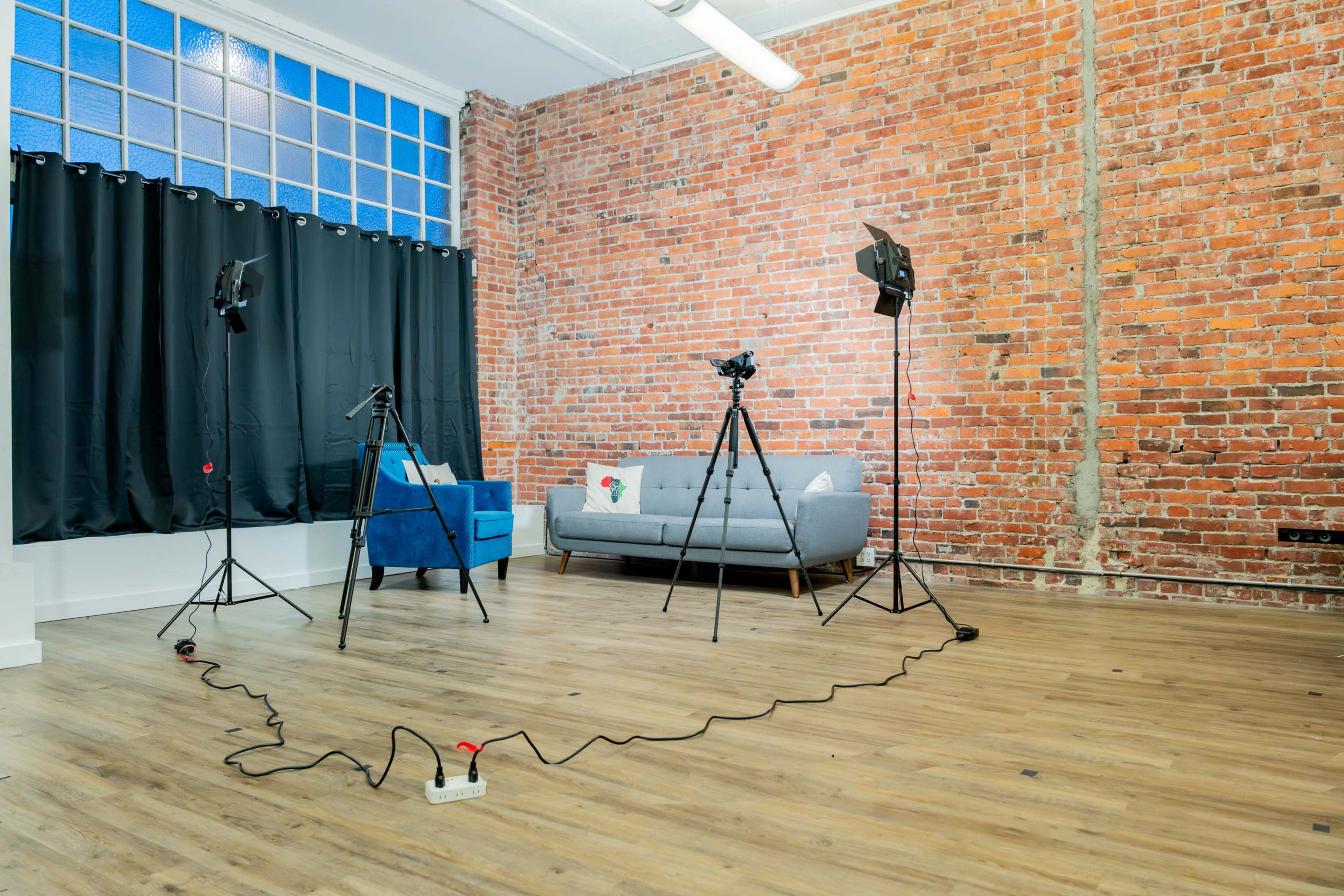 A photo studio features a gray sofa and three tripods with lights set up on a wooden floor against a backdrop of exposed brick walls.
