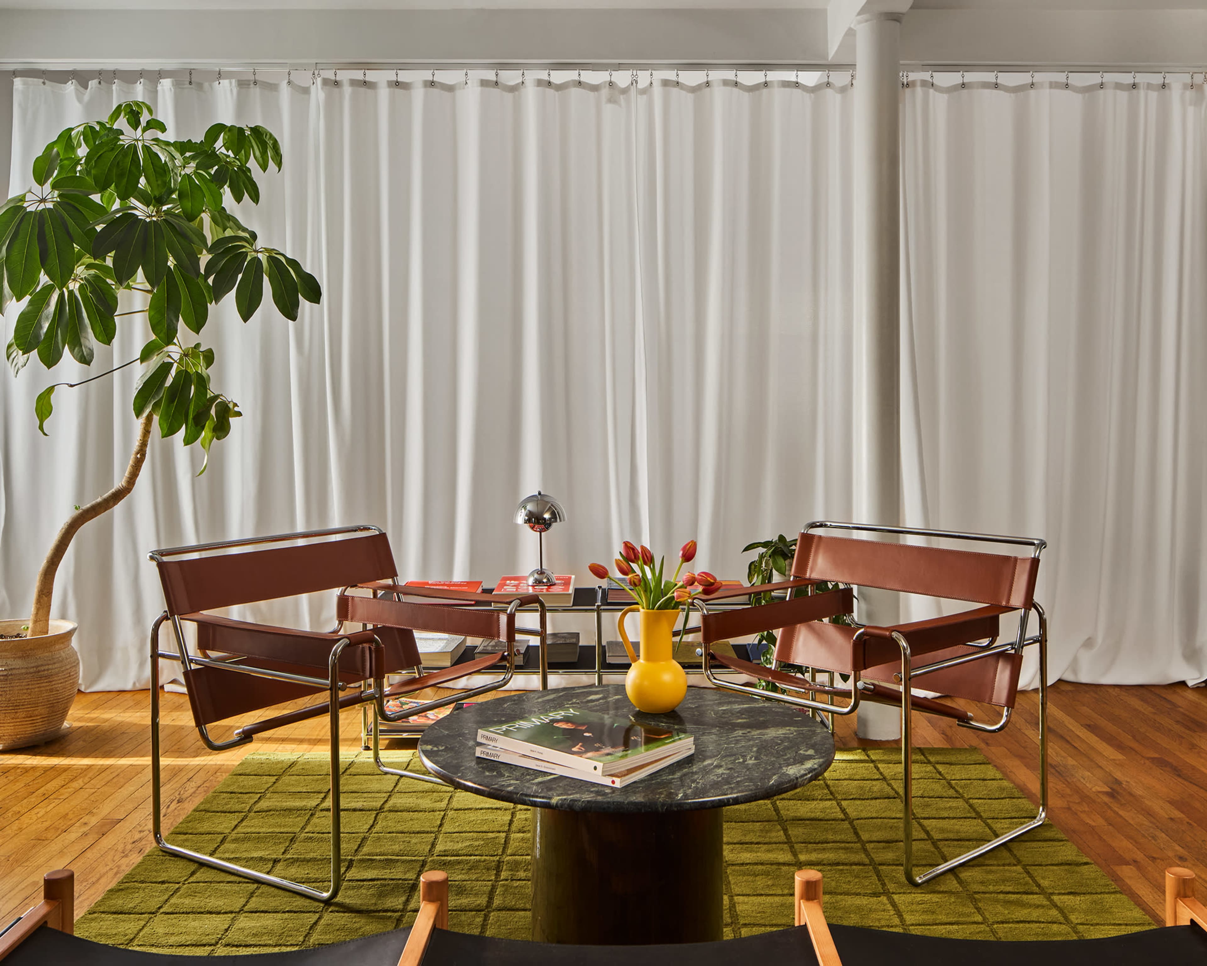 The image shows a modern living area with two brown chairs, a round marble coffee table, and a potted plant, all set against white curtains and a green rug on wooden flooring.