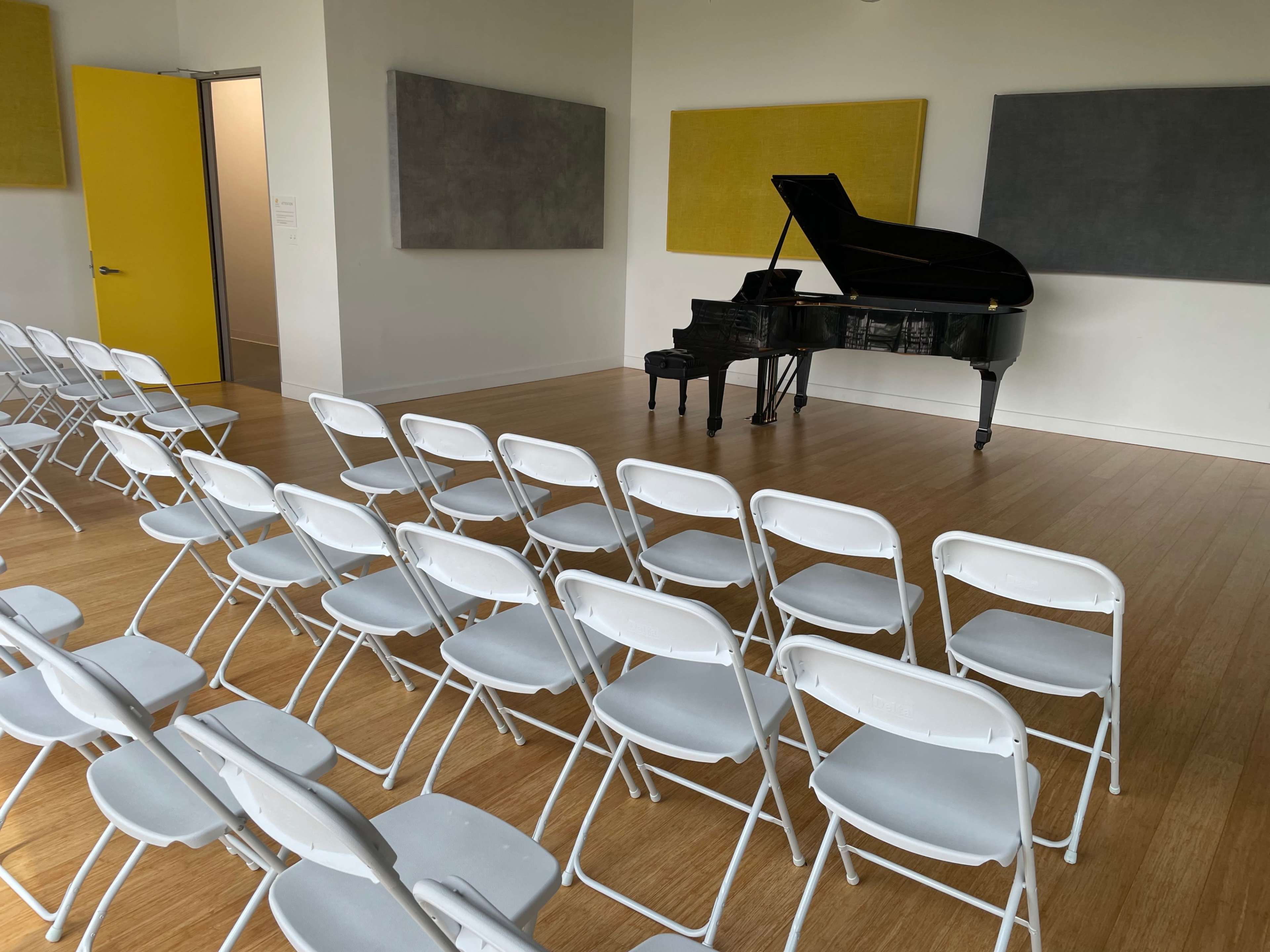 The image shows a room with a grand piano, a small piano, and several rows of white folding chairs set up in front of them.