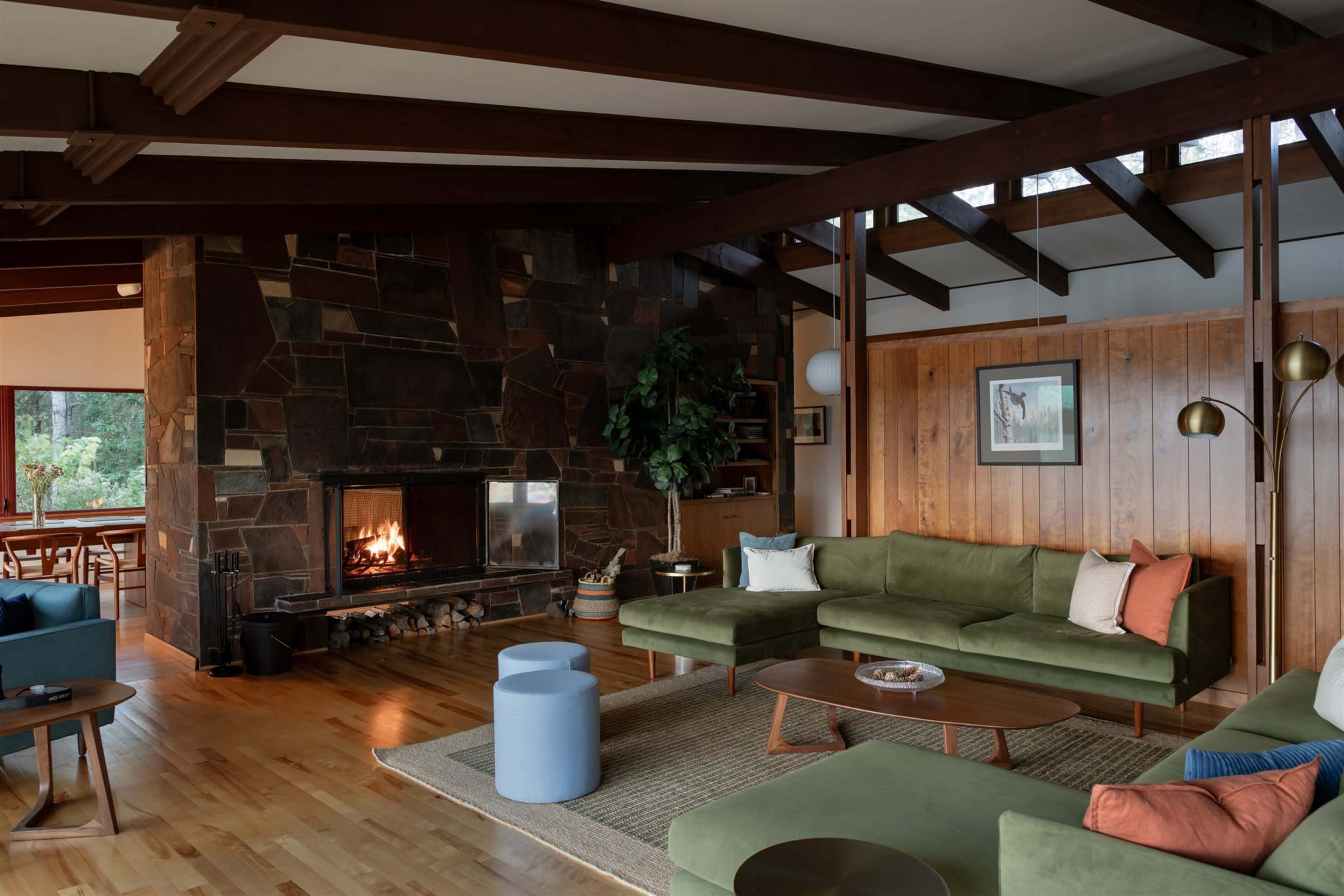 The image shows a modern living room with a stone fireplace, wooden beams, and a green sectional couch, accompanied by a coffee table and an accent chair.