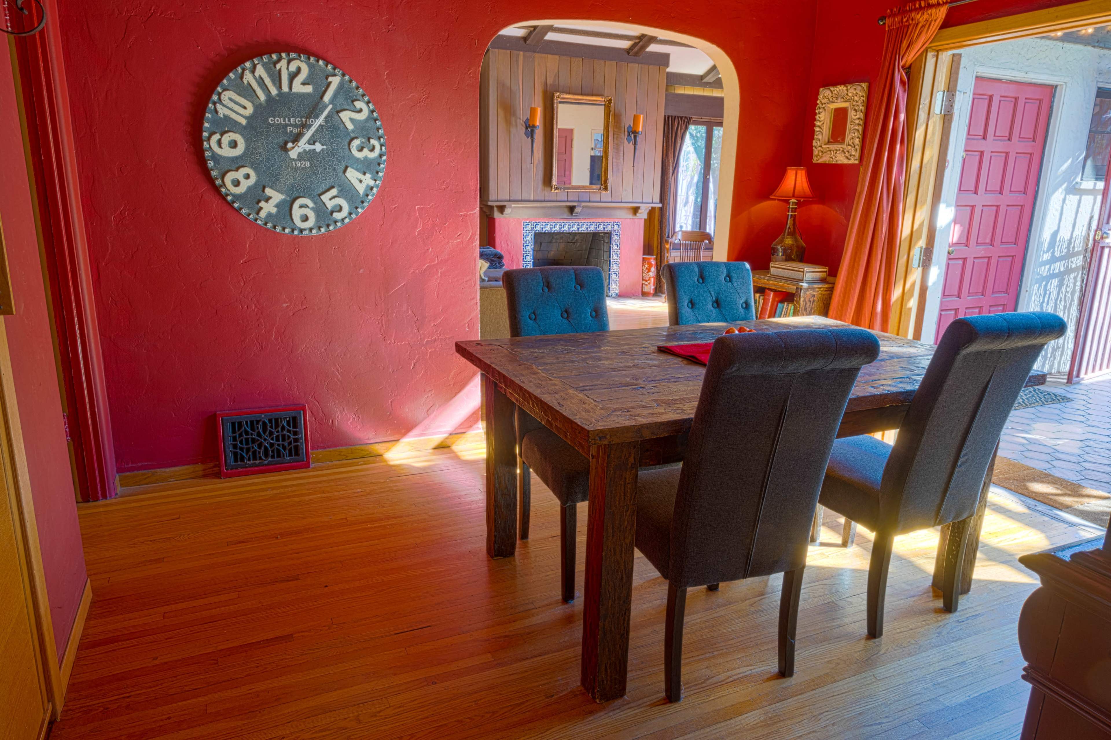 A wooden dining table with upholstered chairs is positioned in a warmly colored room that features a large wall clock and a red door leading outside.