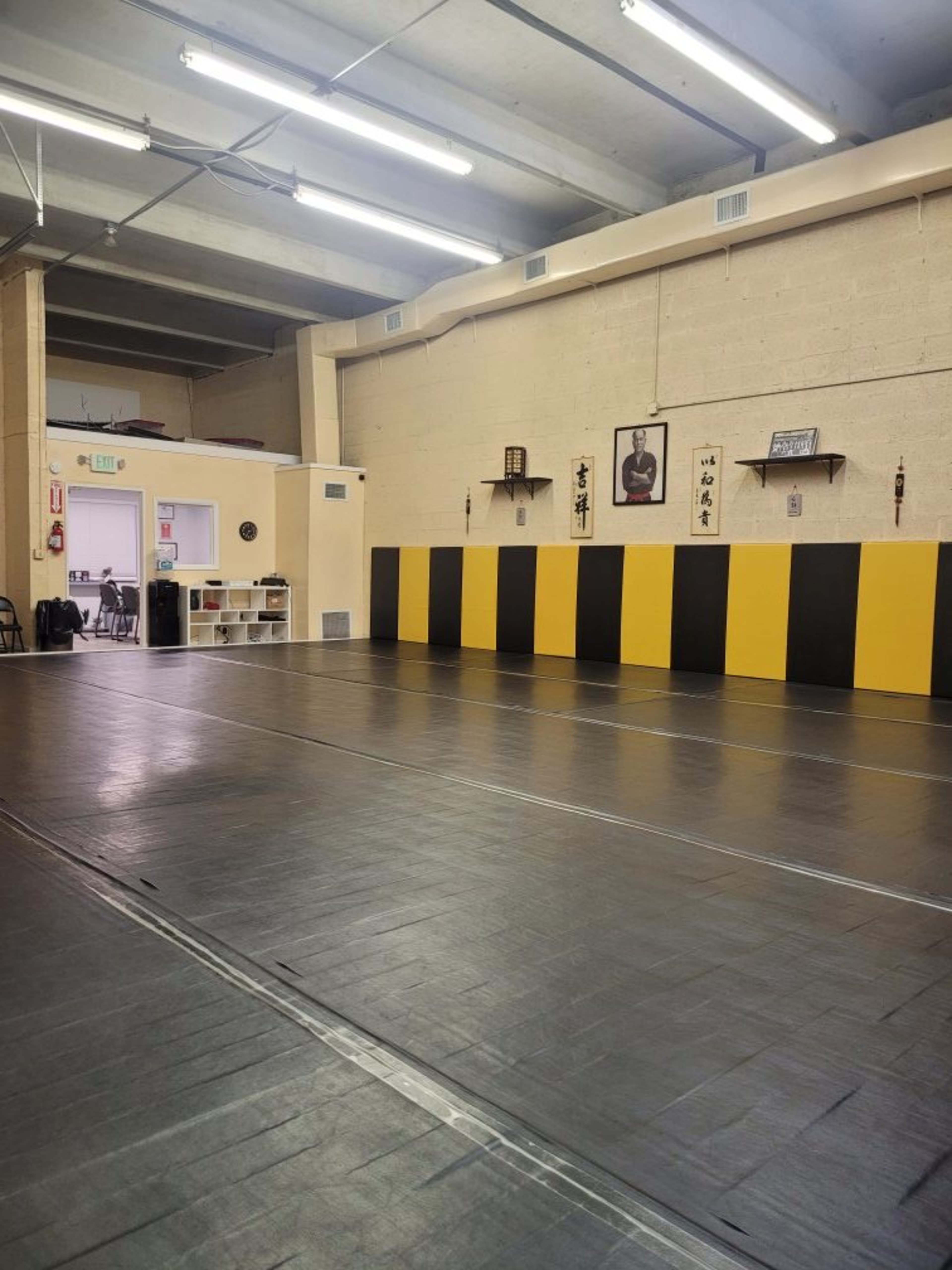The image shows a spacious martial arts studio with black and yellow padded walls, a smooth, dark floor, and various wall decor including traditional martial arts symbols.