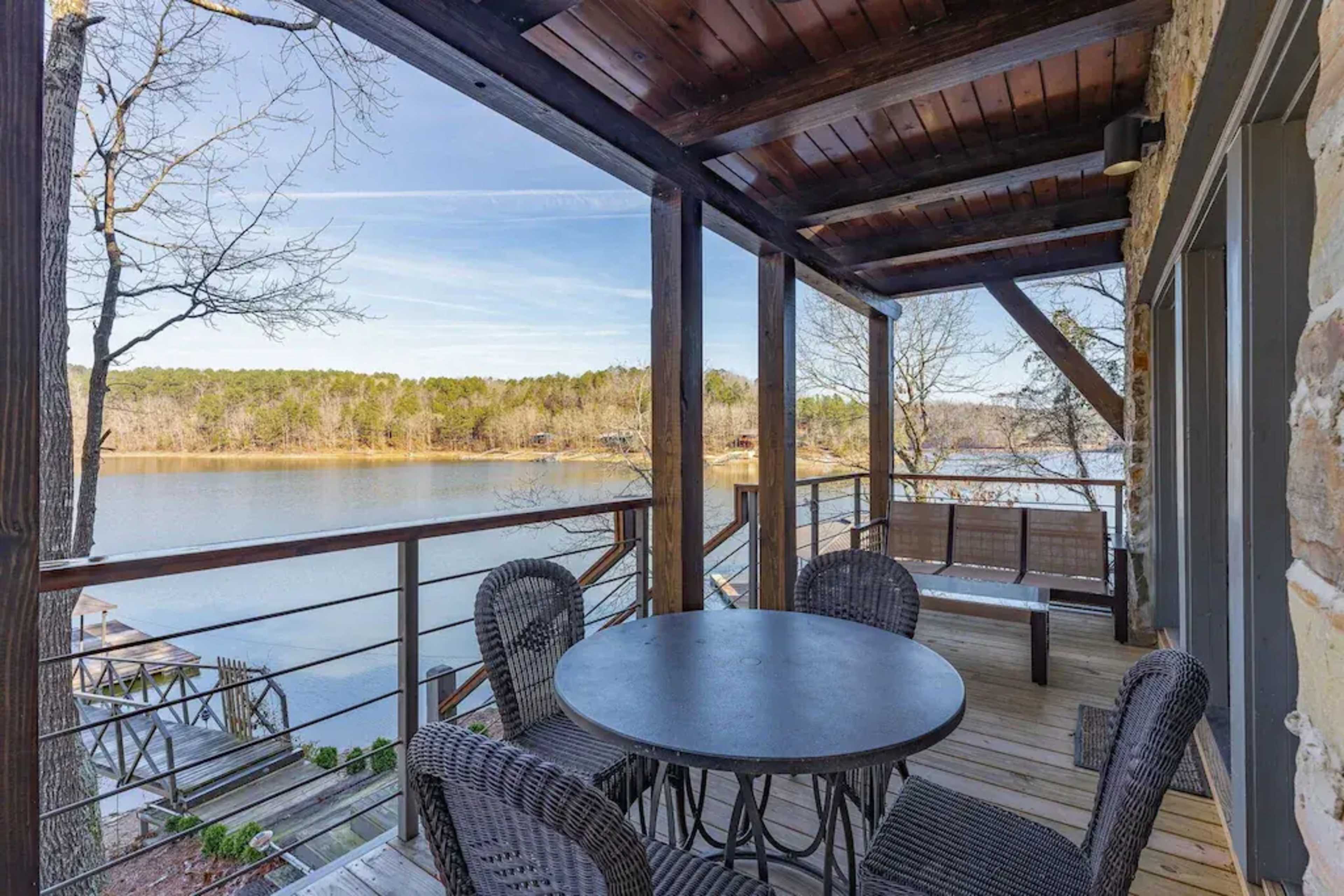 A wooden deck with a circular table and chairs overlooks a calm lake, framed by trees in the background.