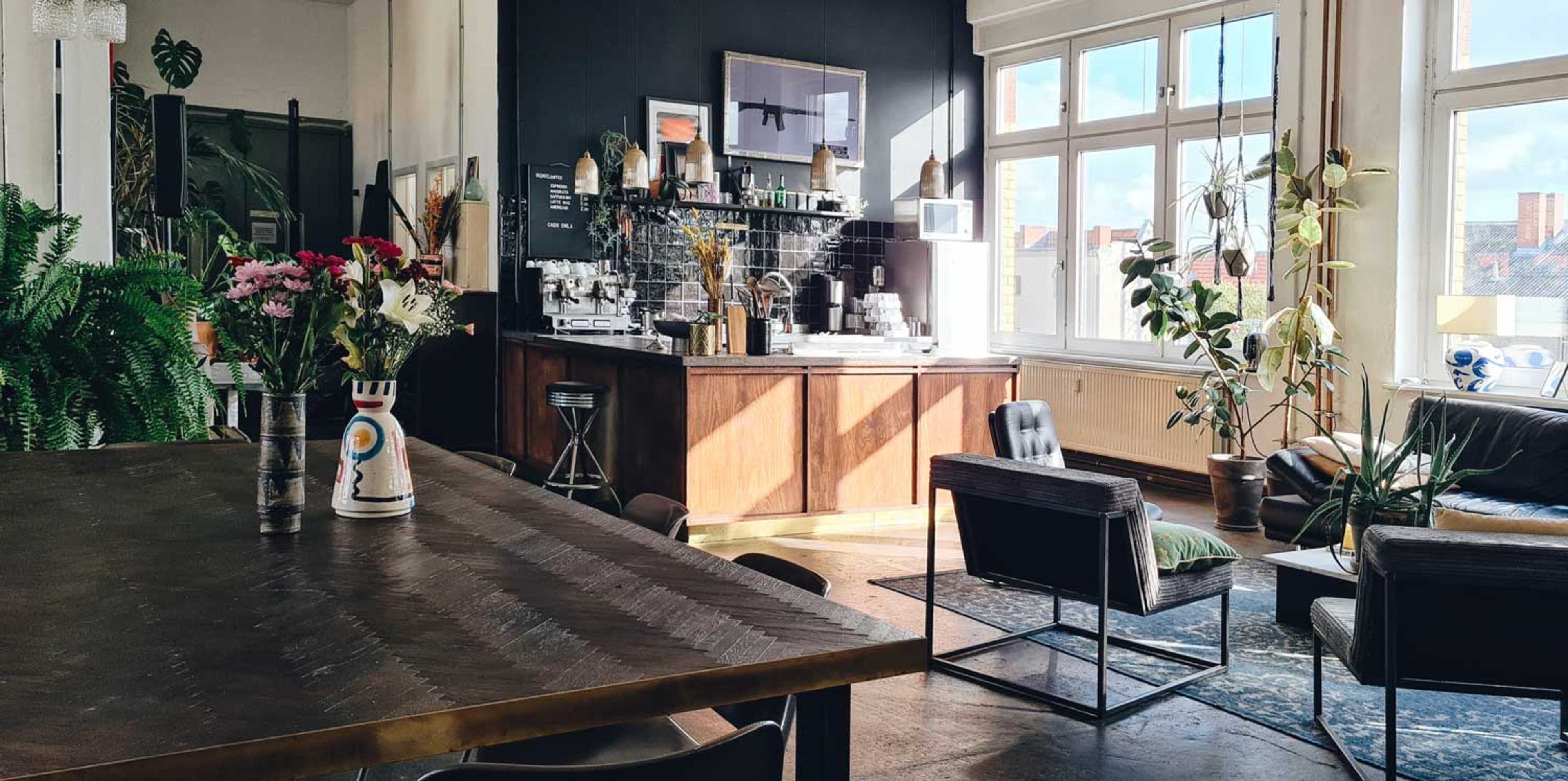The image shows a cozy café interior with a wooden counter, a coffee station, large windows, and various plants decorating the space.