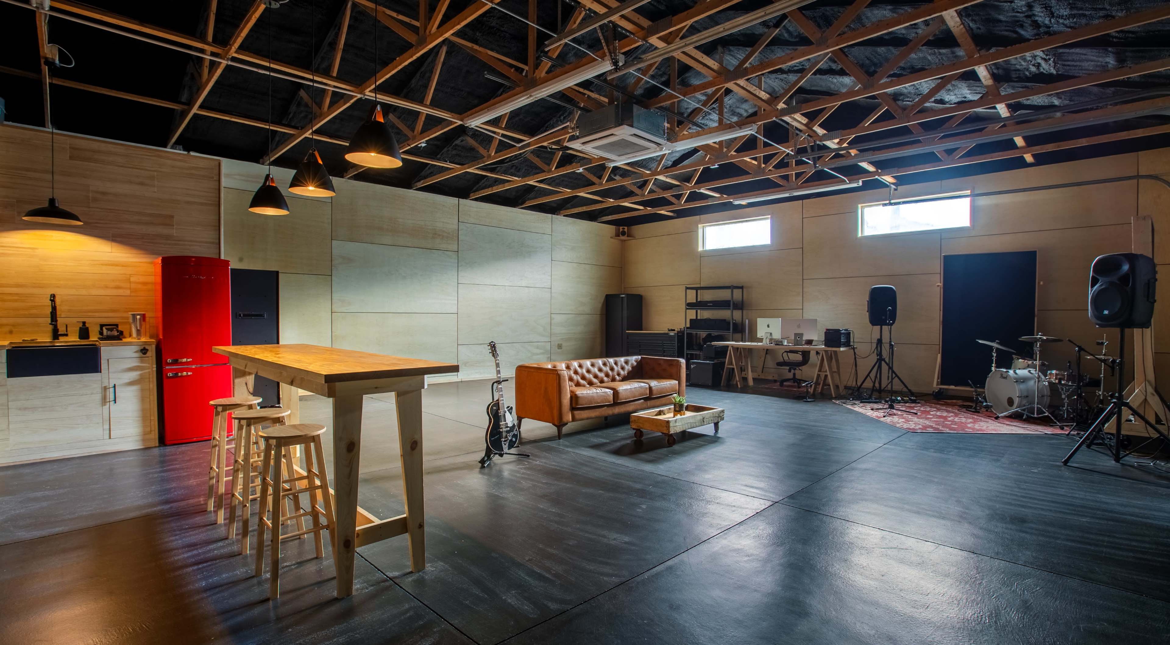 The image shows a spacious studio with wooden walls, a kitchen area, a living space with a leather sofa, and music equipment including a drum set and speakers.