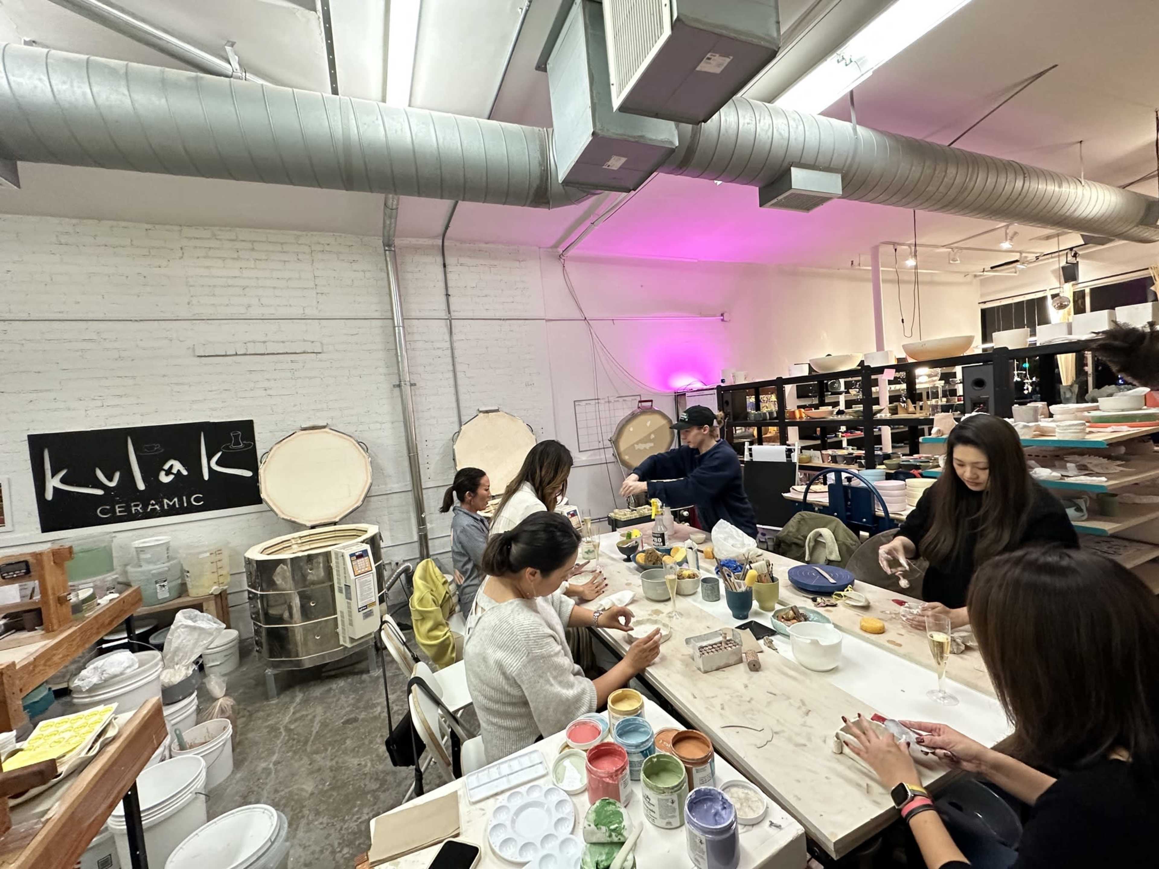 A group of people work on pottery projects at tables in a well-lit ceramics studio featuring several kilns in the background.