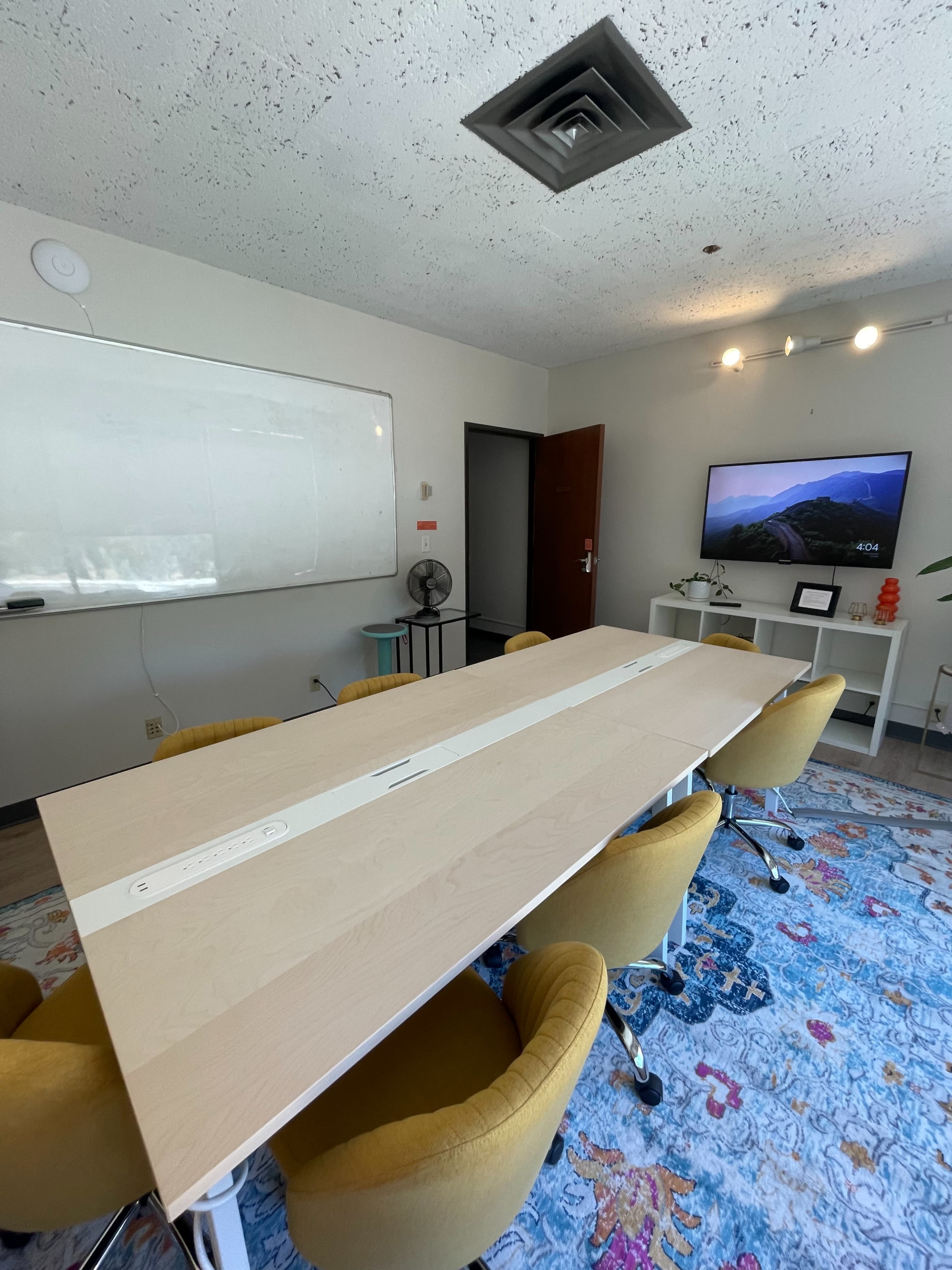 A conference room features a large rectangular table surrounded by yellow chairs, a whiteboard on one wall, and a television on another displaying a landscape.