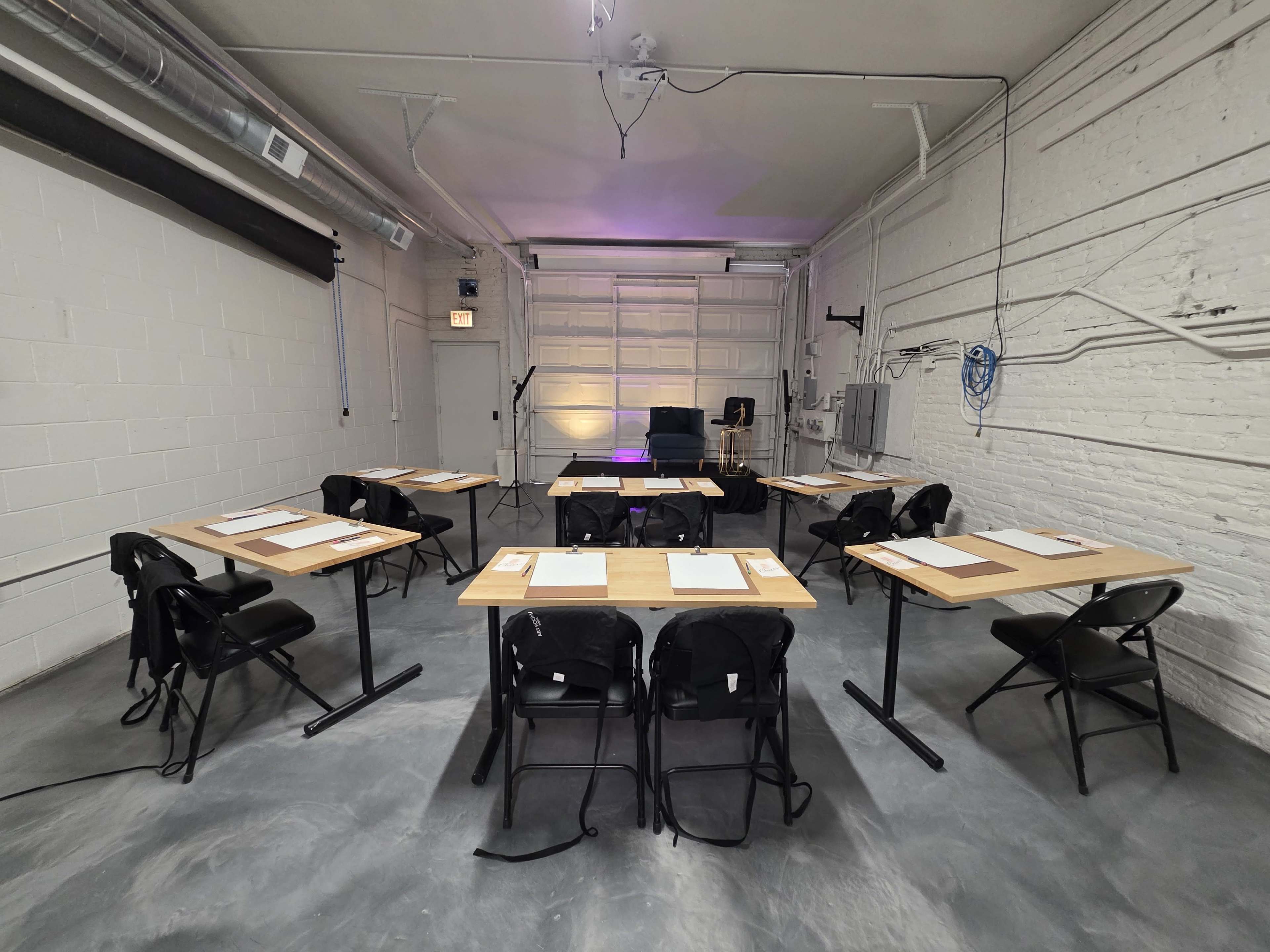 The image shows a blank room with several tables arranged in a U-shape, each equipped with sheets of paper and writing materials, in a space with exposed pipes and a garage door.