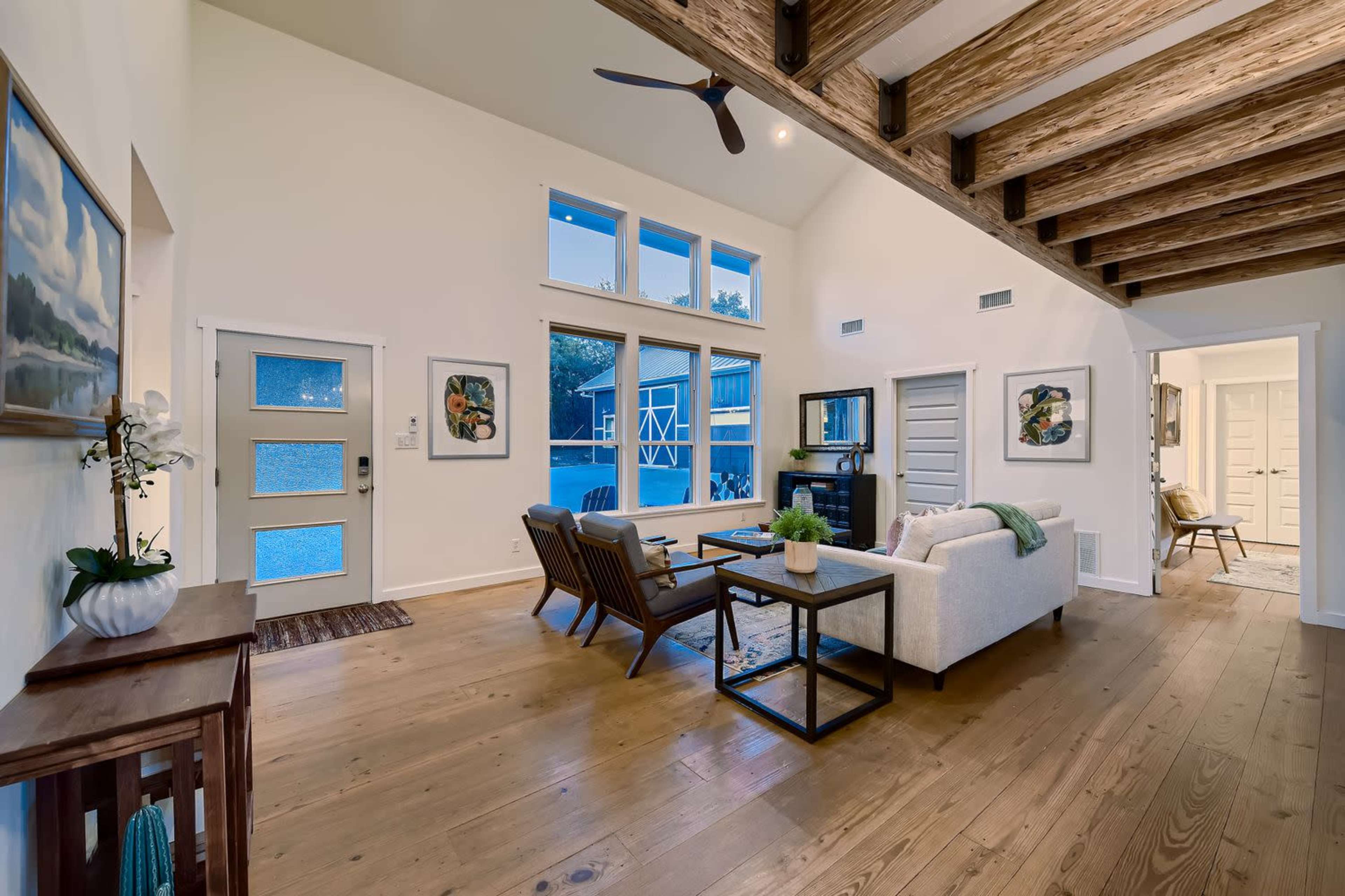 The image shows a modern living room with high ceilings, wooden beams, and large windows overlooking a backyard pool.