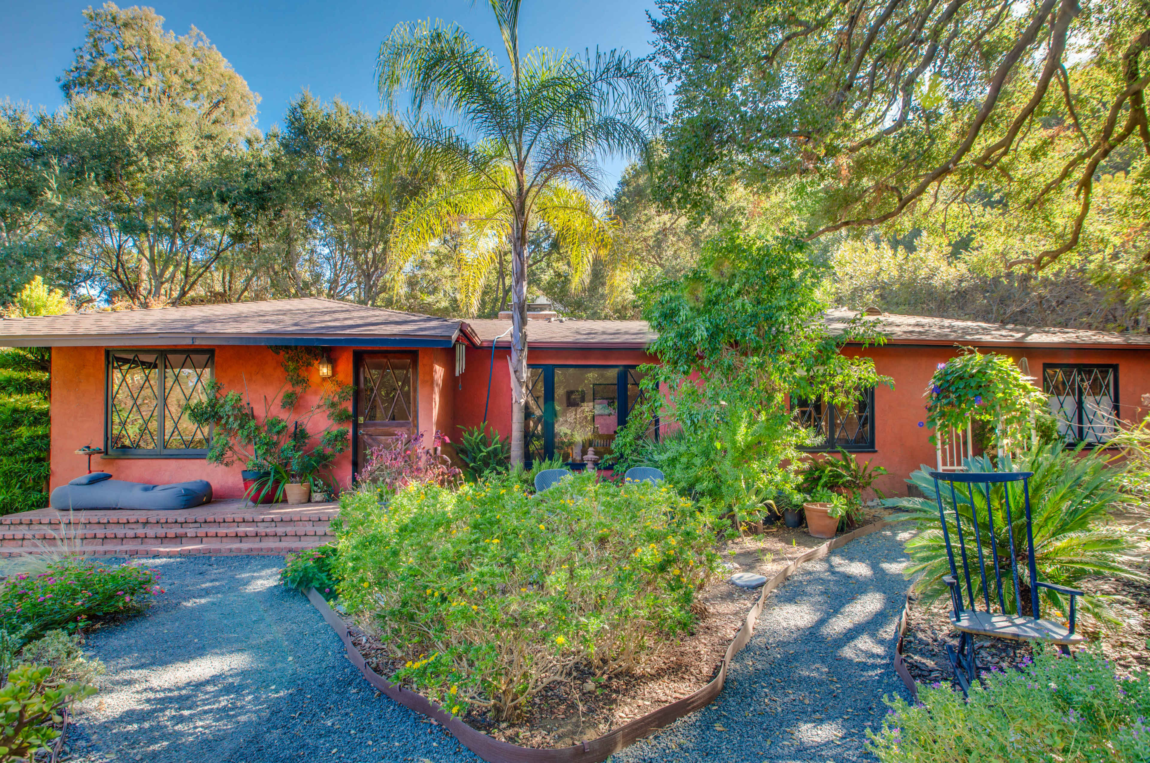 The image shows a vibrant, single-story home surrounded by lush greenery and a pathway lined with plants.