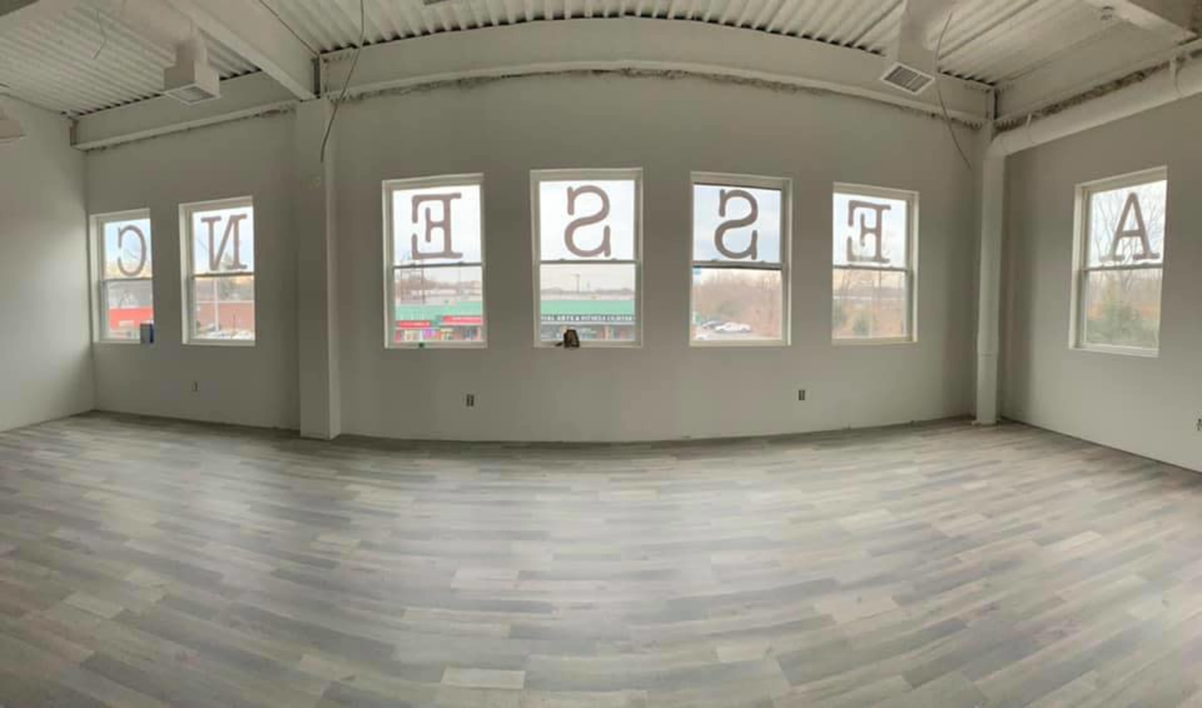 The image shows an empty room with gray wooden flooring and large windows featuring the word "ESSENCE" prominently displayed.