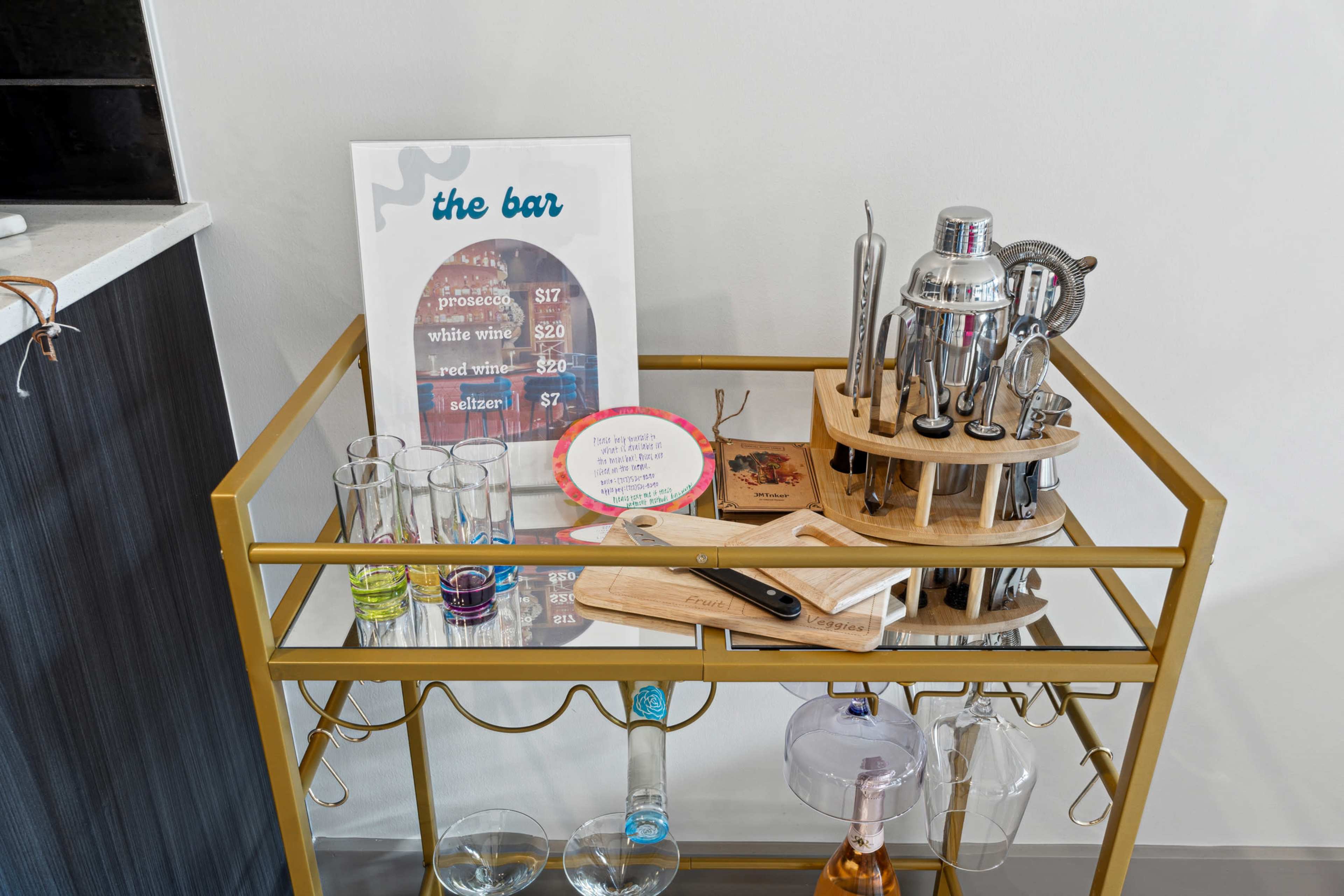 A stylish bar cart displays a cocktail shaker set, various glasses, and a menu of drink prices beside a cutting board.