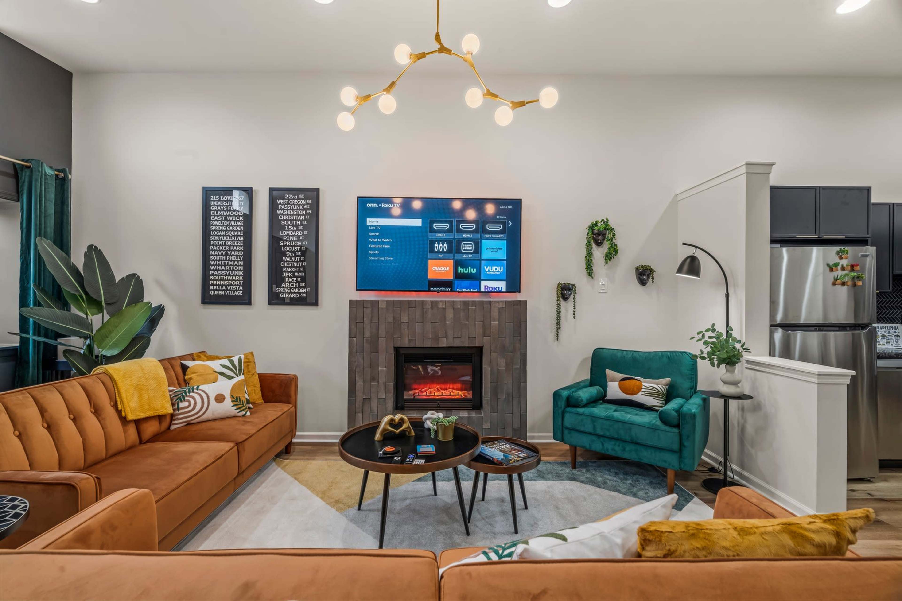 A modern living room features a sectional sofa, a decorative fireplace, a television on the wall, and plants in the corners.