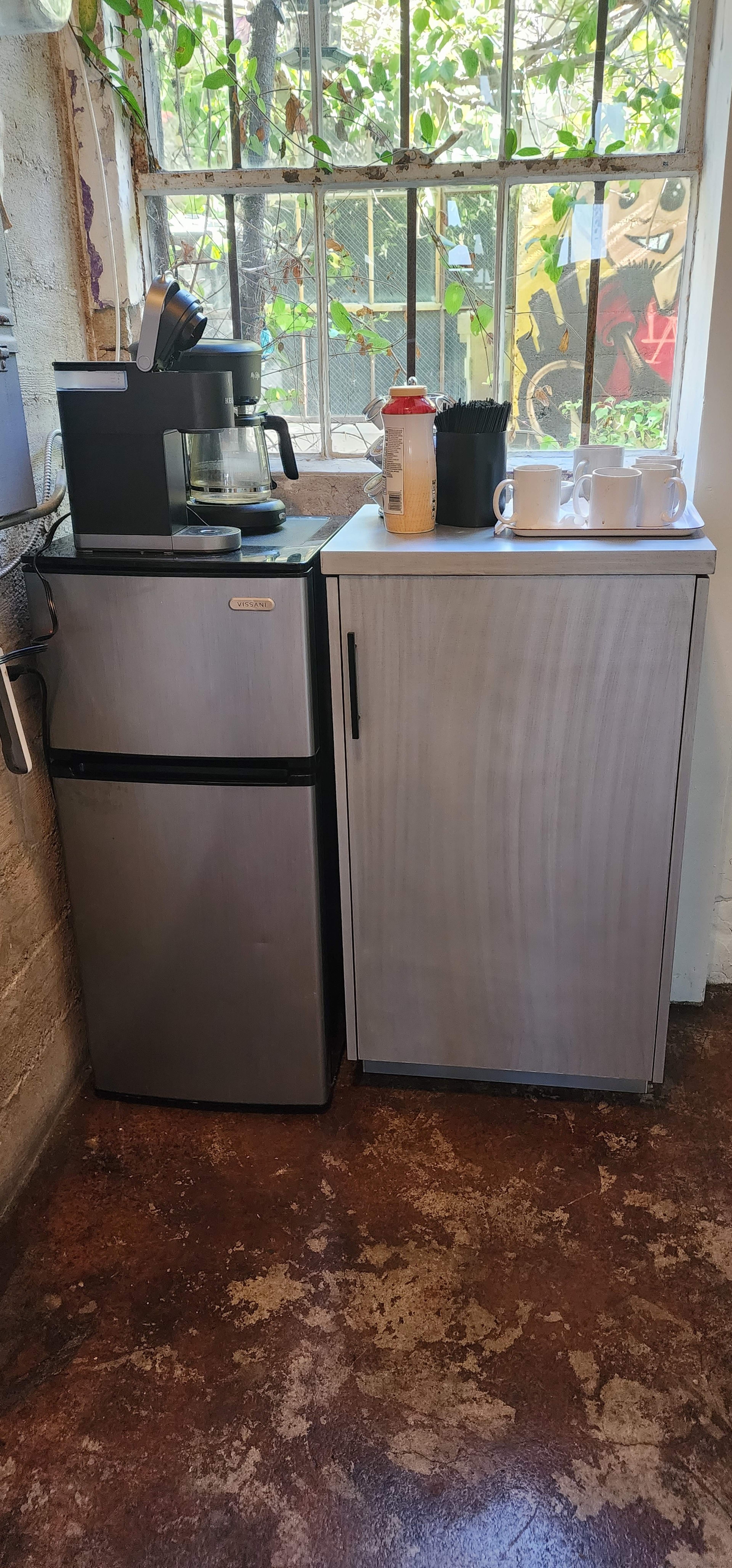 A small kitchenette area with a coffee maker on a counter next to two compact refrigerators under a window with greenery outside.