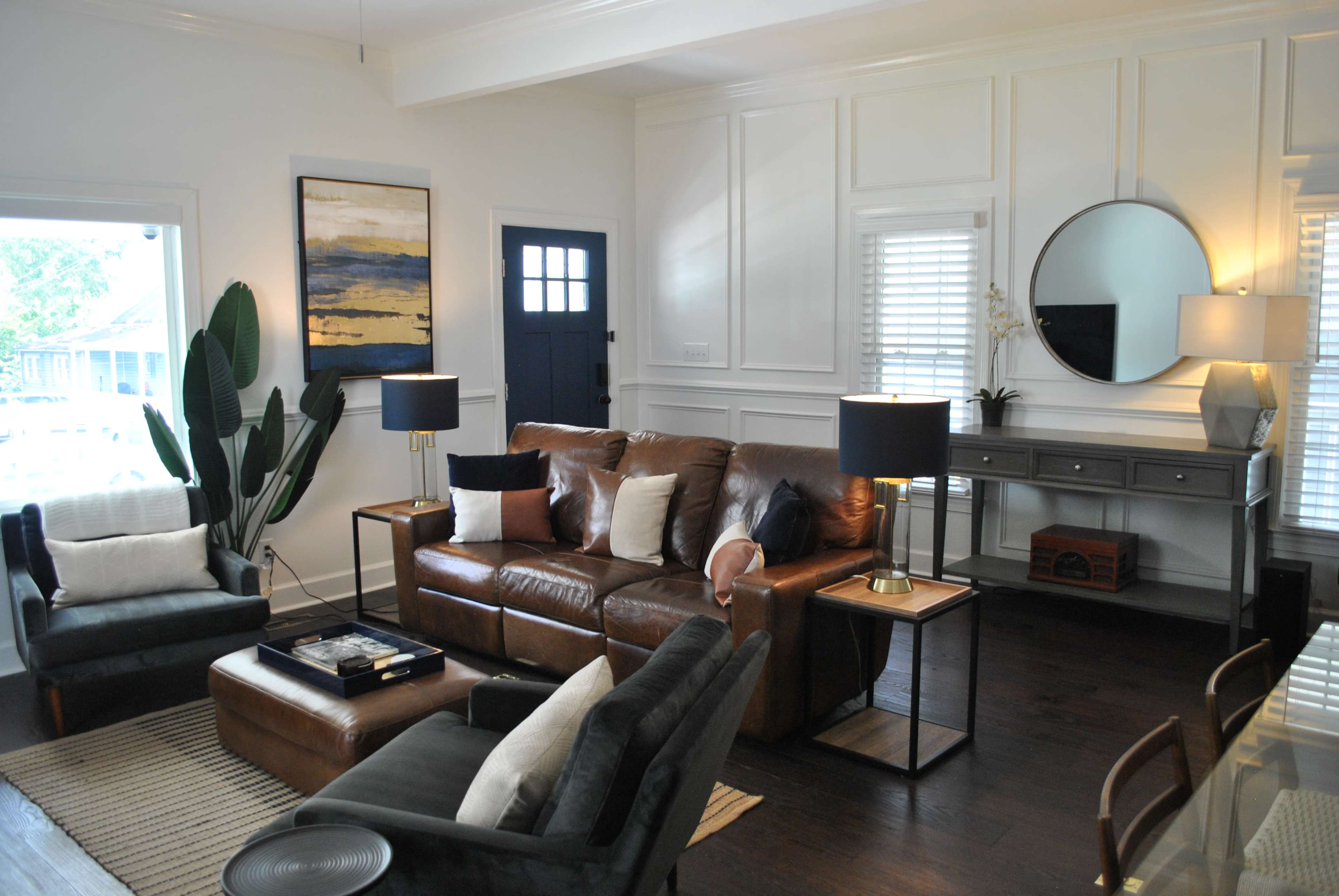 The living room features a brown leather sofa, two dark chairs, a wooden side table, and decorative plants against a backdrop of white shiplap walls and large windows.
