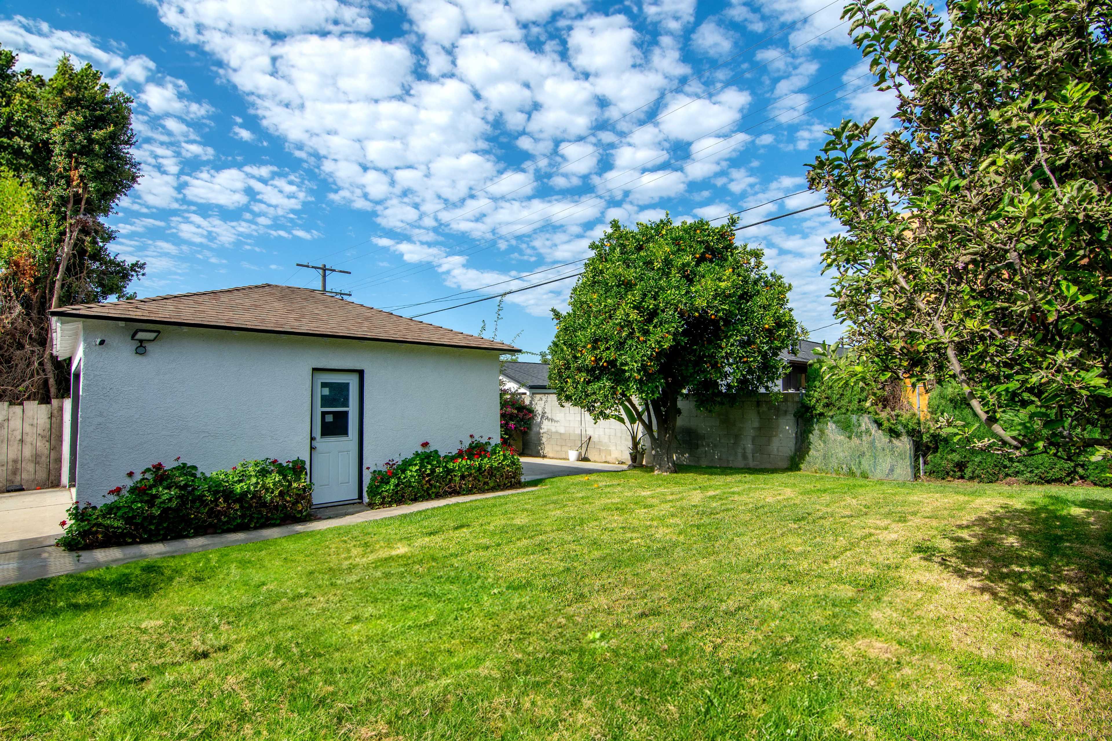 The image shows a well-maintained backyard featuring a small white building, lush green grass, and several trees under a partly cloudy sky.